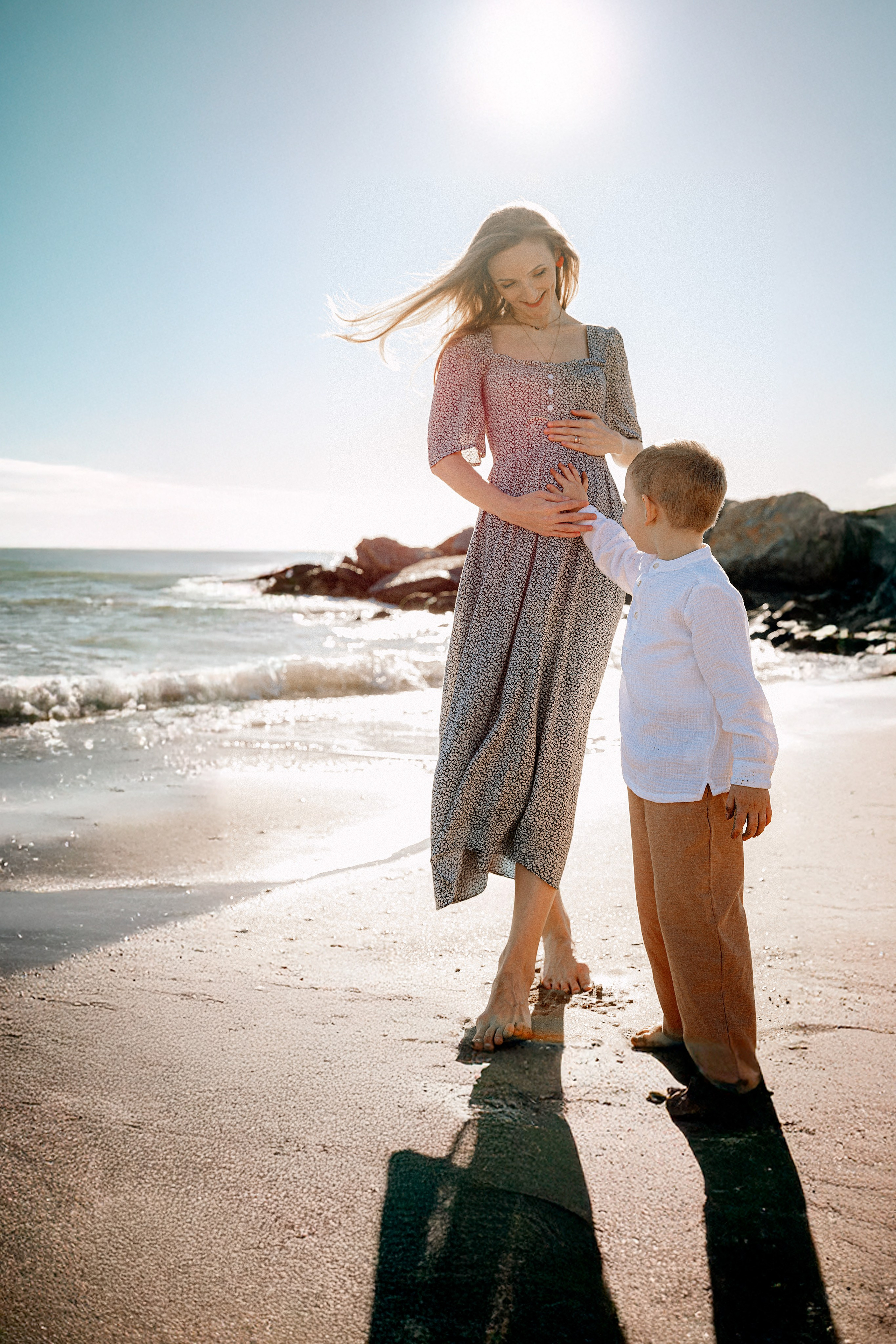 Sesión de fotos familiar en València, España, que captura un delicado momento junto al mar de una madre y su hijo pequeño de pie descalzos frente al mar Mediterráneo, mientras el niño toca suavemente su vientre, fotografiado con luz natural suave, una conexión emocional profunda y una atmósfera costera tranquila — ideal para fotografía familiar, sesiones de maternidad y familia, retratos familiares lifestyle y sesiones emotivas en la playa en València y en toda España.