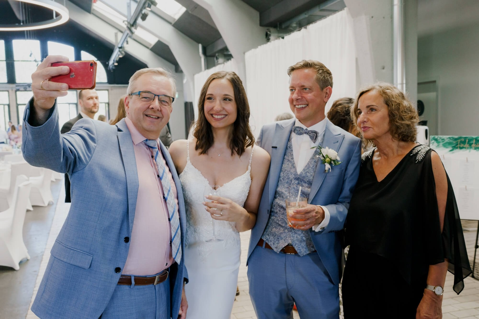 A guest taking a selfie with the newlyweds and another guest in a bright industrial-style reception hall