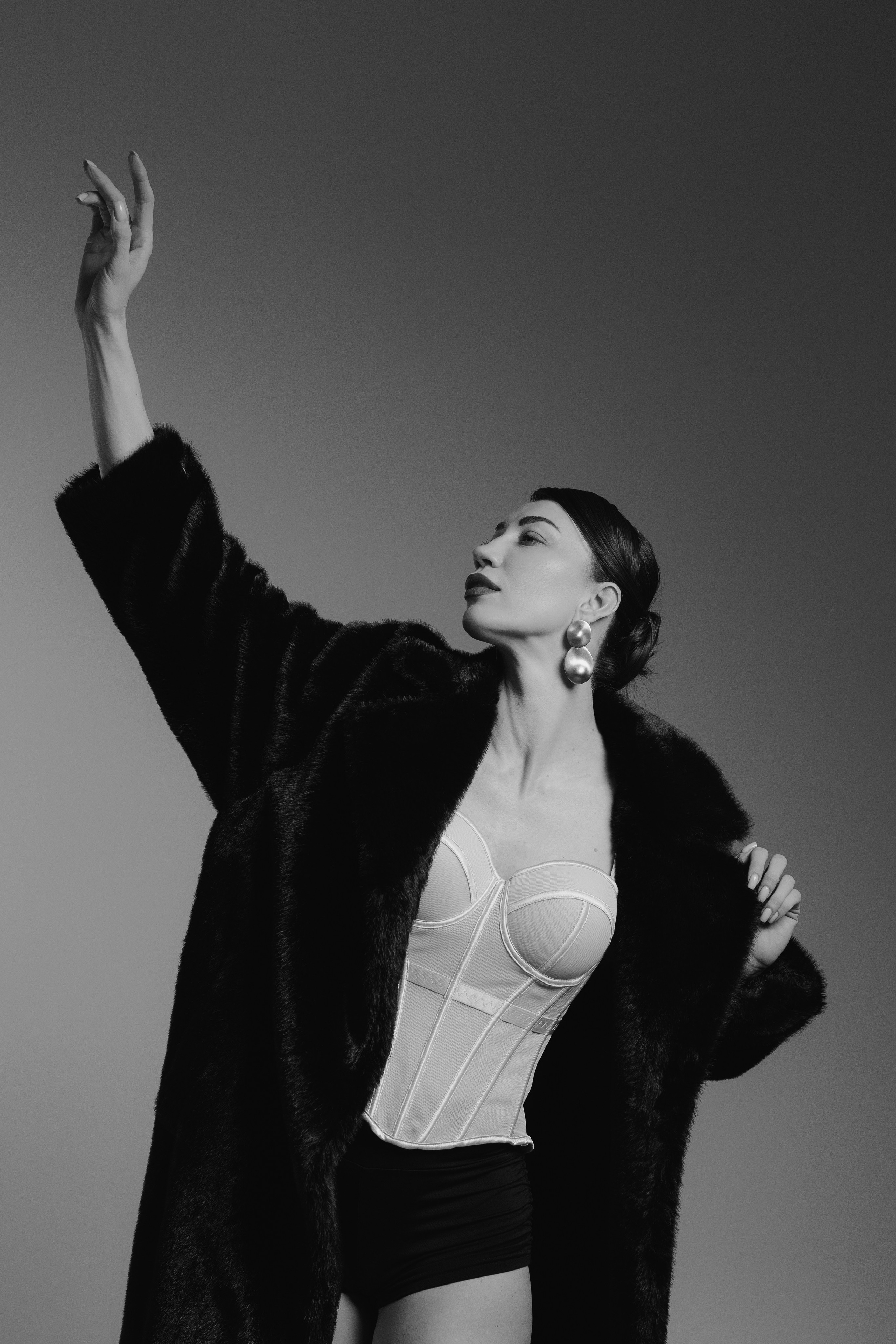 Dramatic editorial studio portrait of a woman in a black fur coat raising her arm in expressive pose with cinematic lighting, photographed in Leipzig, Germany. Драматичный редакционный студийный портрет девушки в чёрной шубе с поднятой рукой в выразительной позе и кинематографичном освещении, снятый в Лейпциге, Германия.