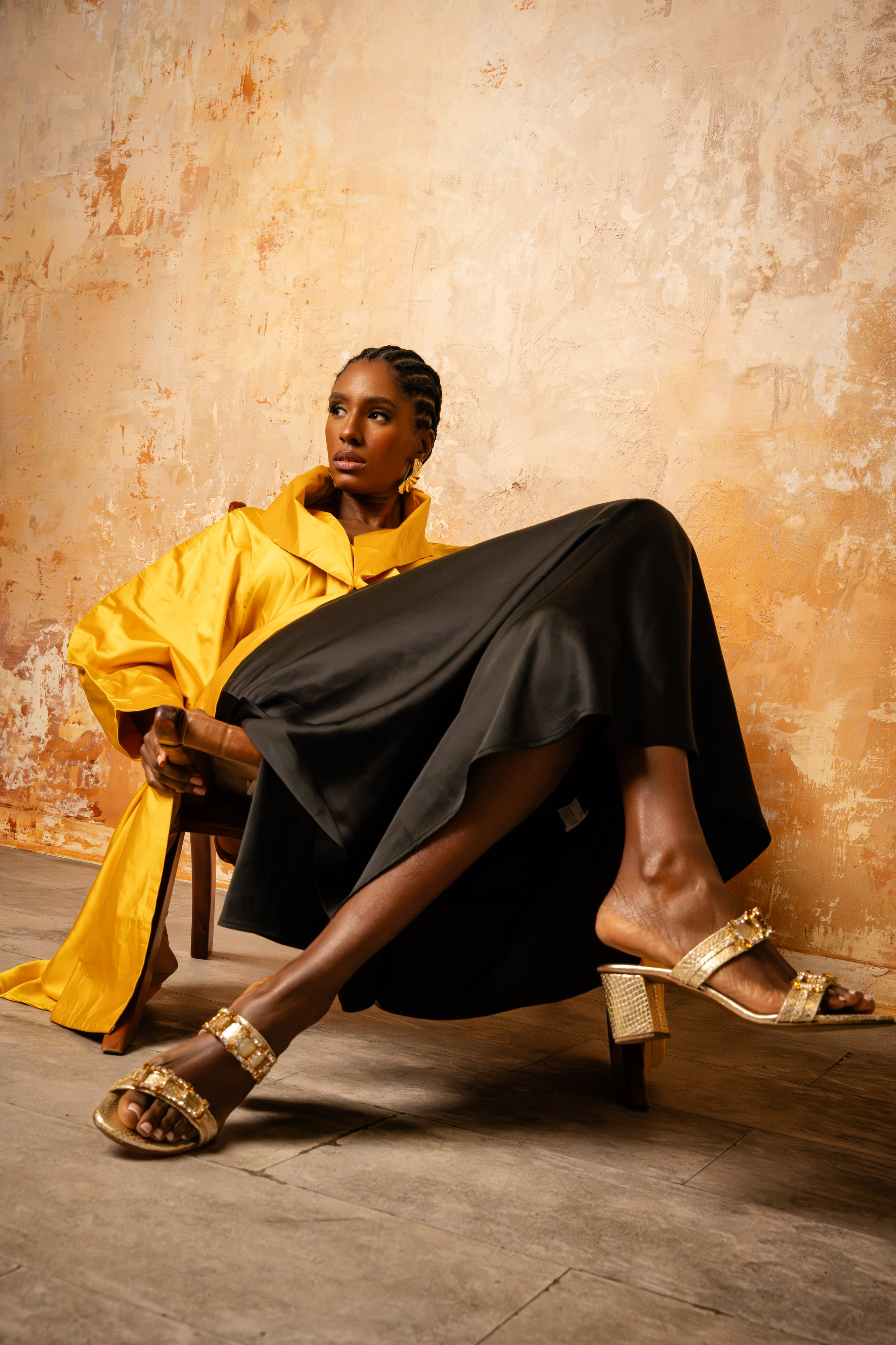 Editorial fashion portrait of a seated woman wearing yellow and black styling against a warm textured backdrop, photographed in a polished studio setting.