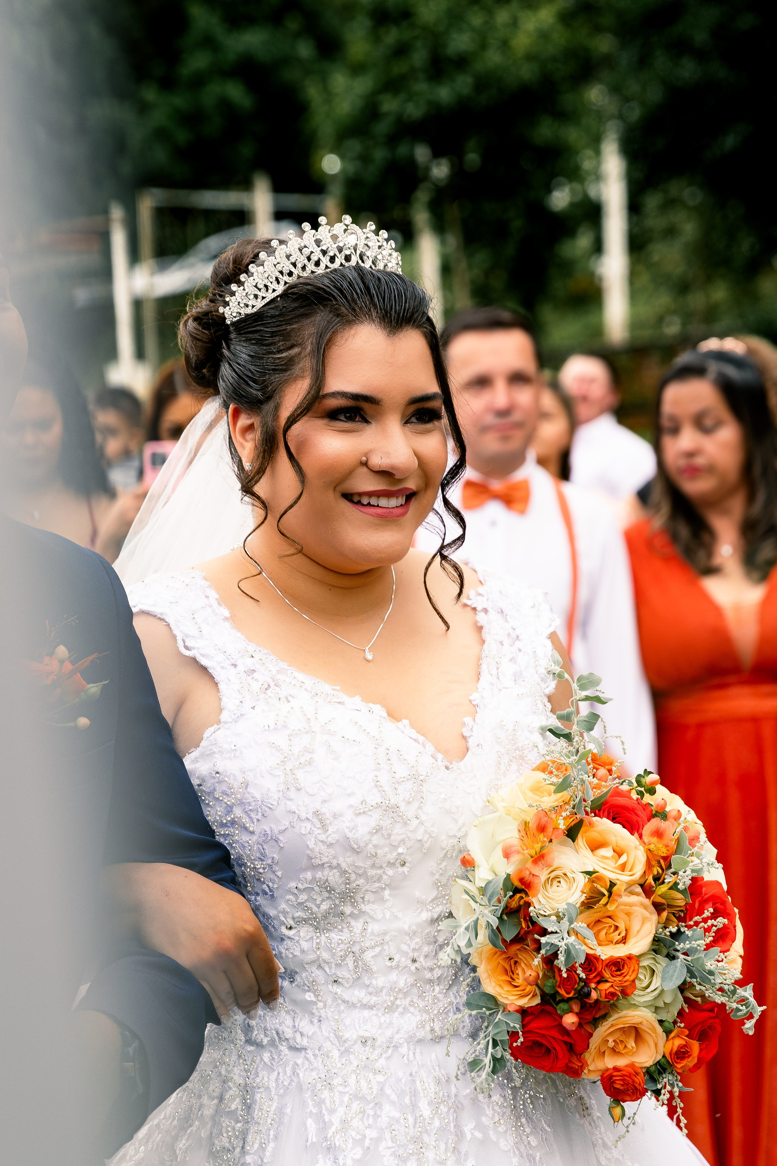 Noiva com tiara e buquê colorido sorrindo suavemente durante a cerimônia, convidados assistindo.