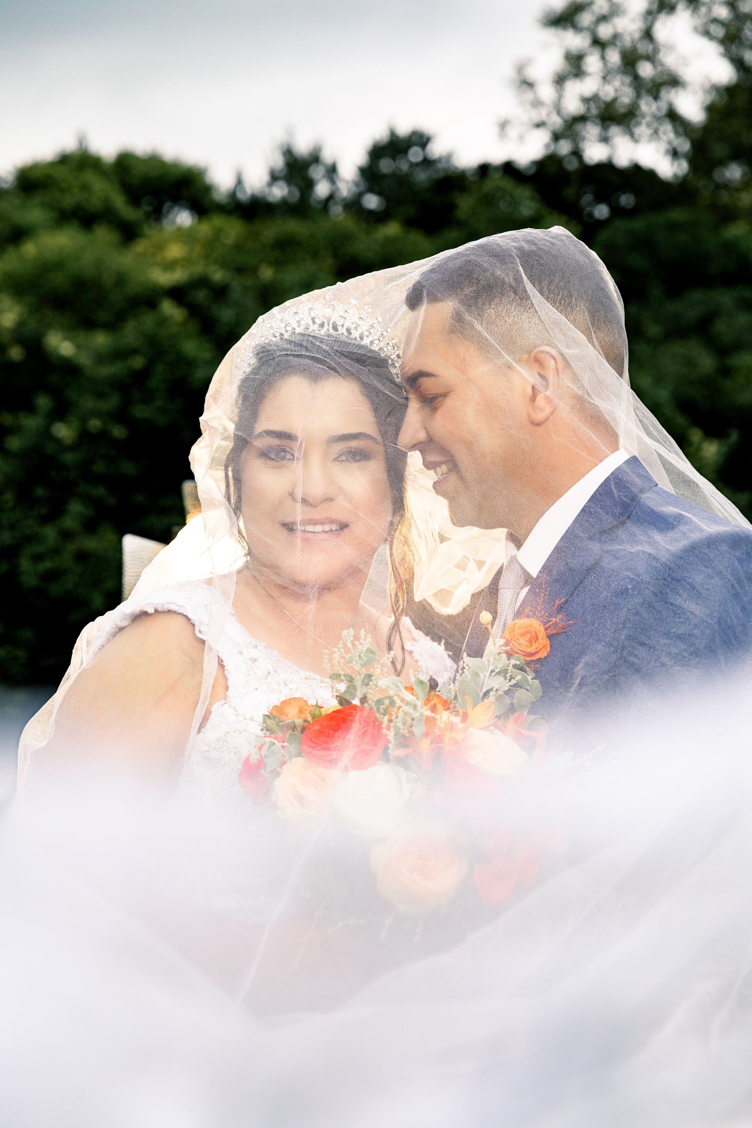 Noiva e noivo sorrindo juntos sob o véu com fundo verde natural, luz suave e conexão visível.