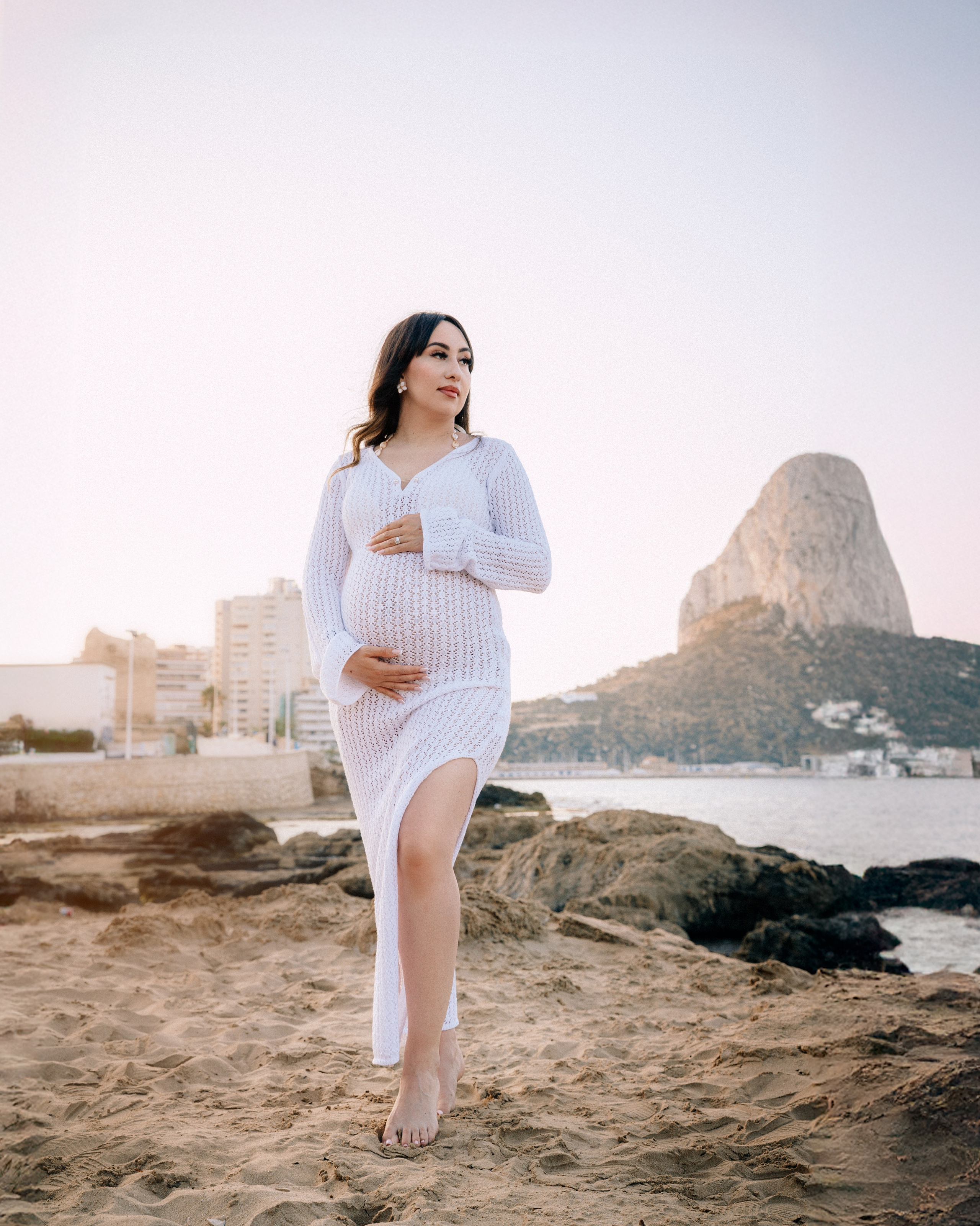 Elegante sesión de fotos de embarazo en Calpe, España — futura madre con vestido blanco posando descalza frente al mar con el Peñón de Ifach al fondo. Inspiración ideal para sesiones fotográficas escénicas de embarazo en la Costa Blanca.