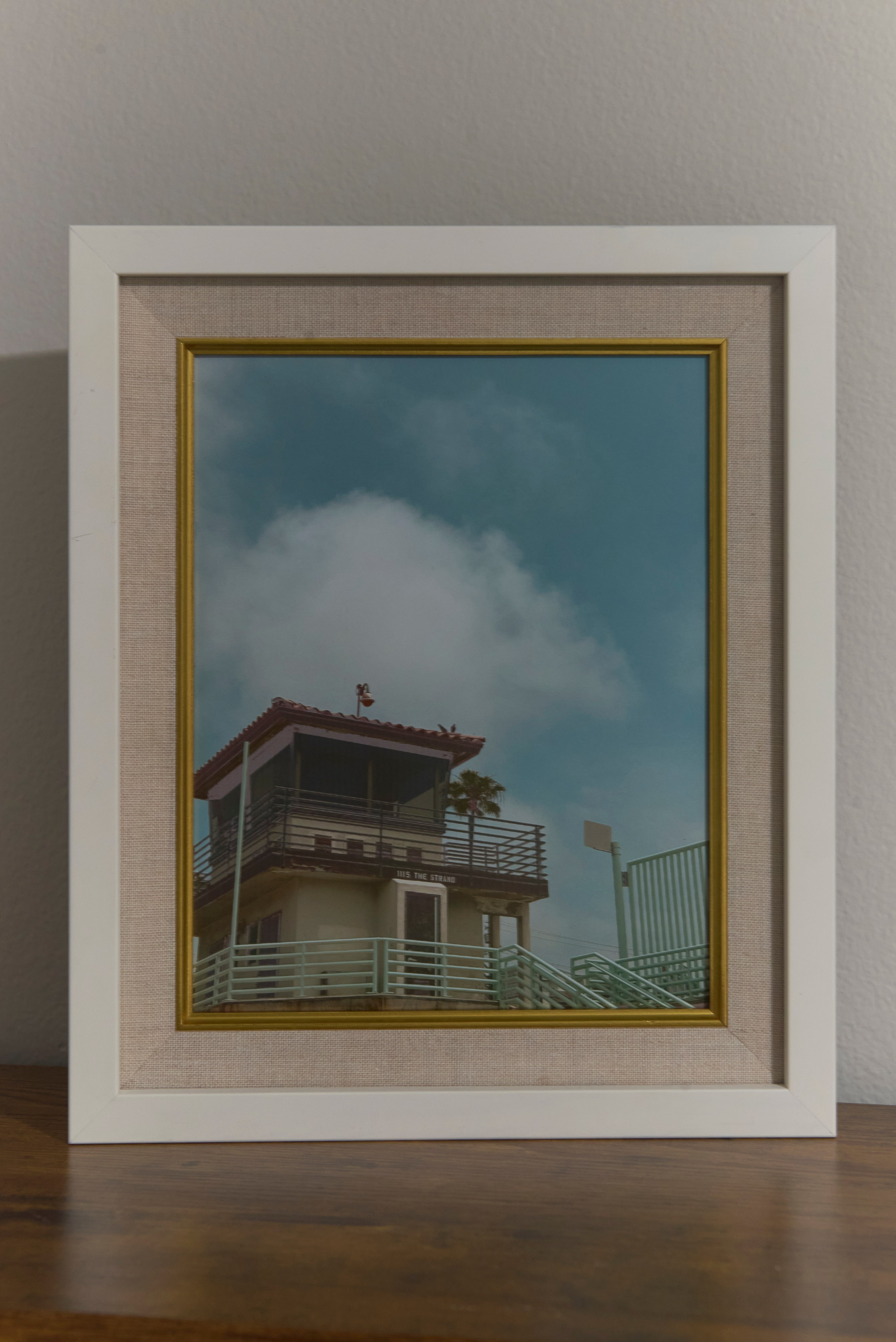 A framed photograph of the Manhattan Beach lifeguard tower with its red roof, balcony railings, and soft cloudy sky in the background.
