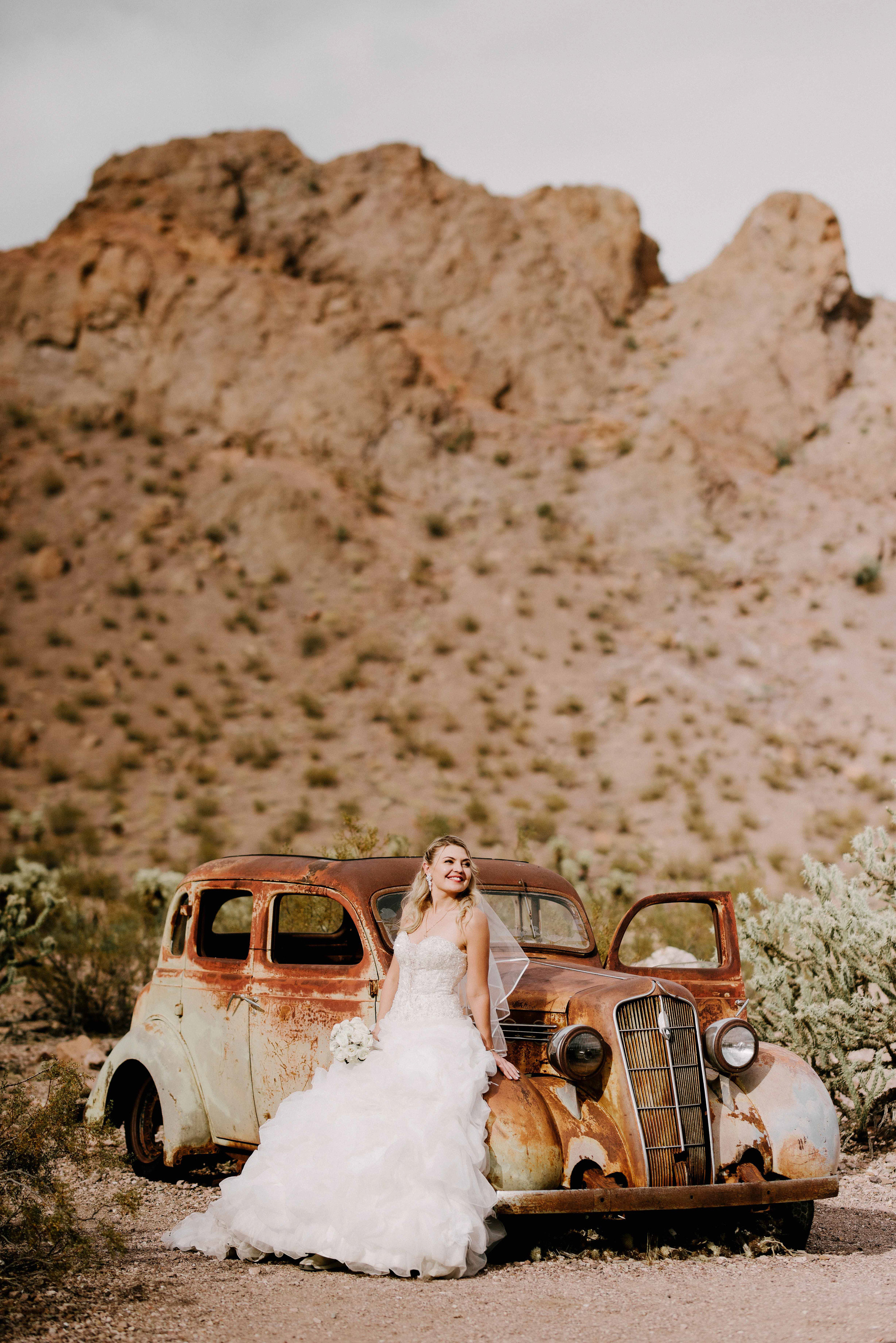 a bride and groom pose in front of a rusty car