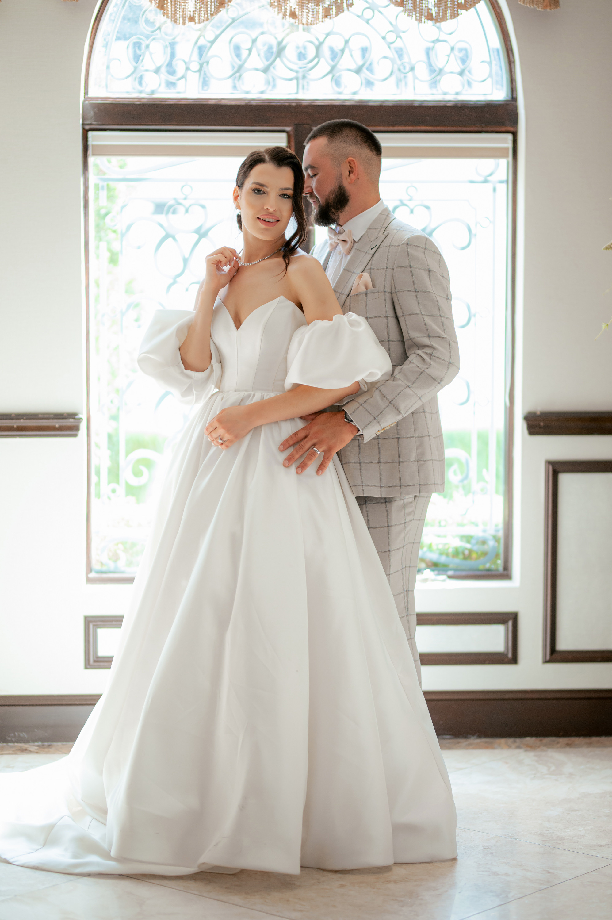 a bride and groom pose for a photo in front of a window
