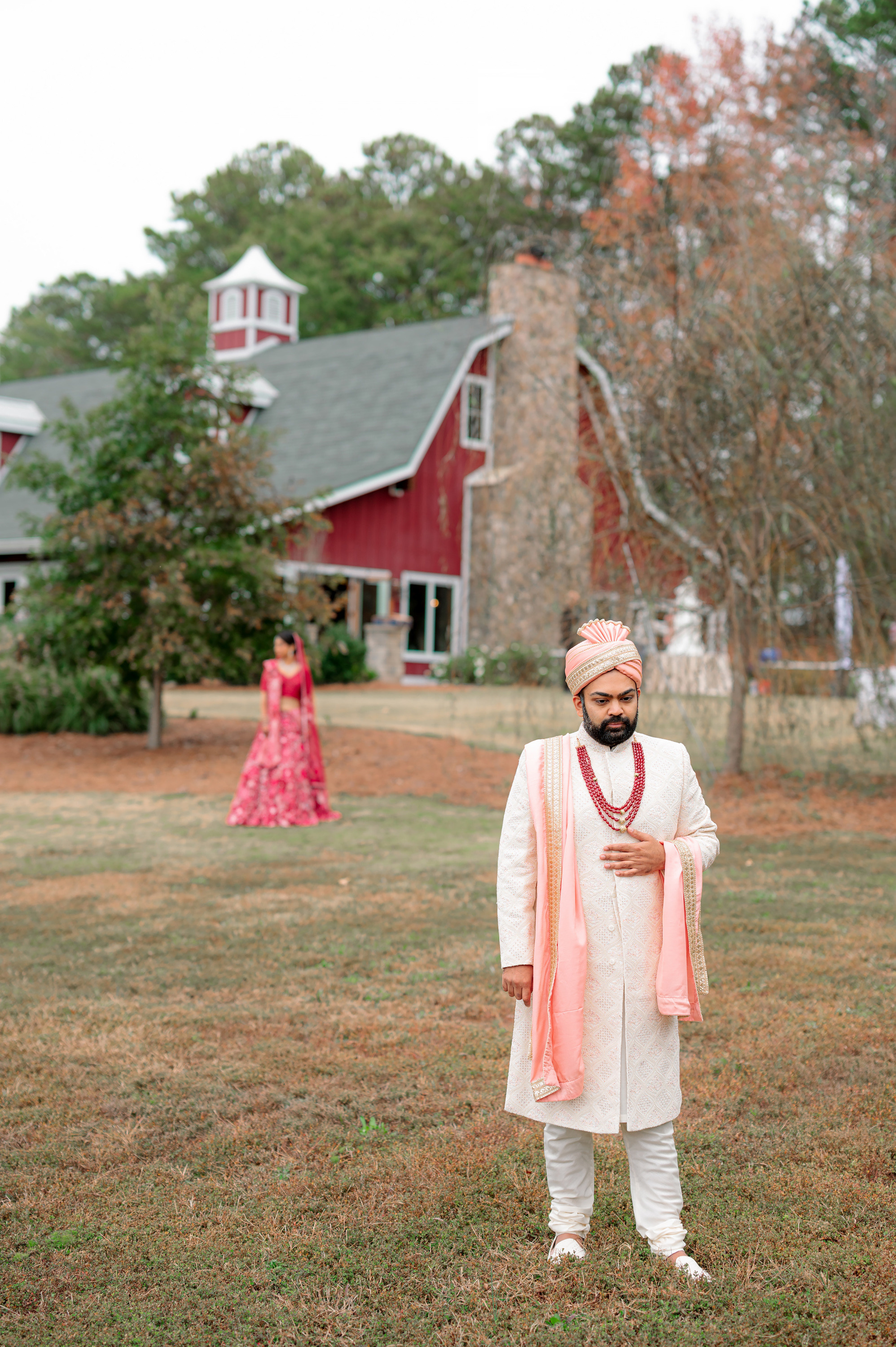 a man in a white suit and pink scarf standing in front of a red barn