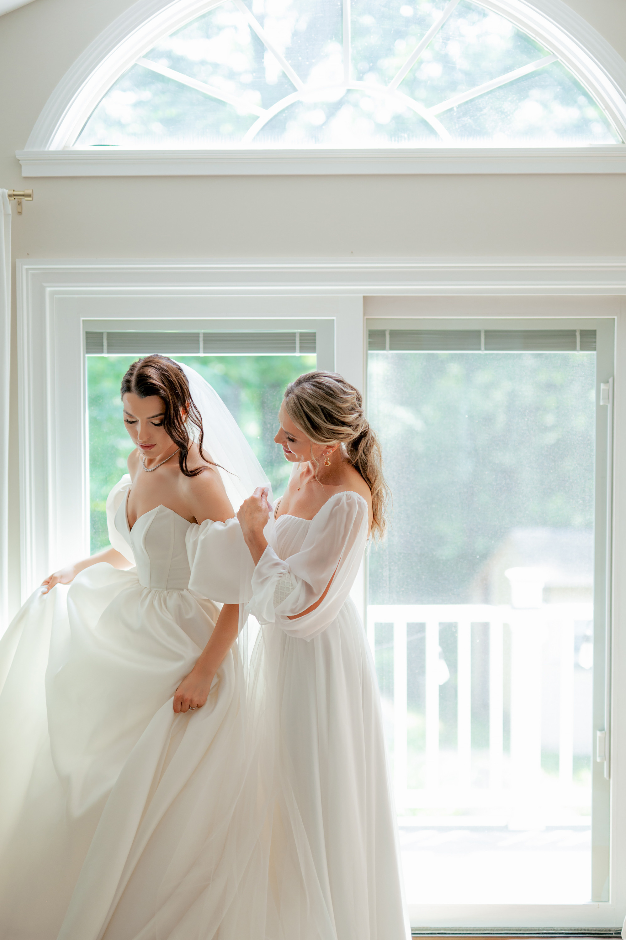 two brides in white dresses standing in front of a window
