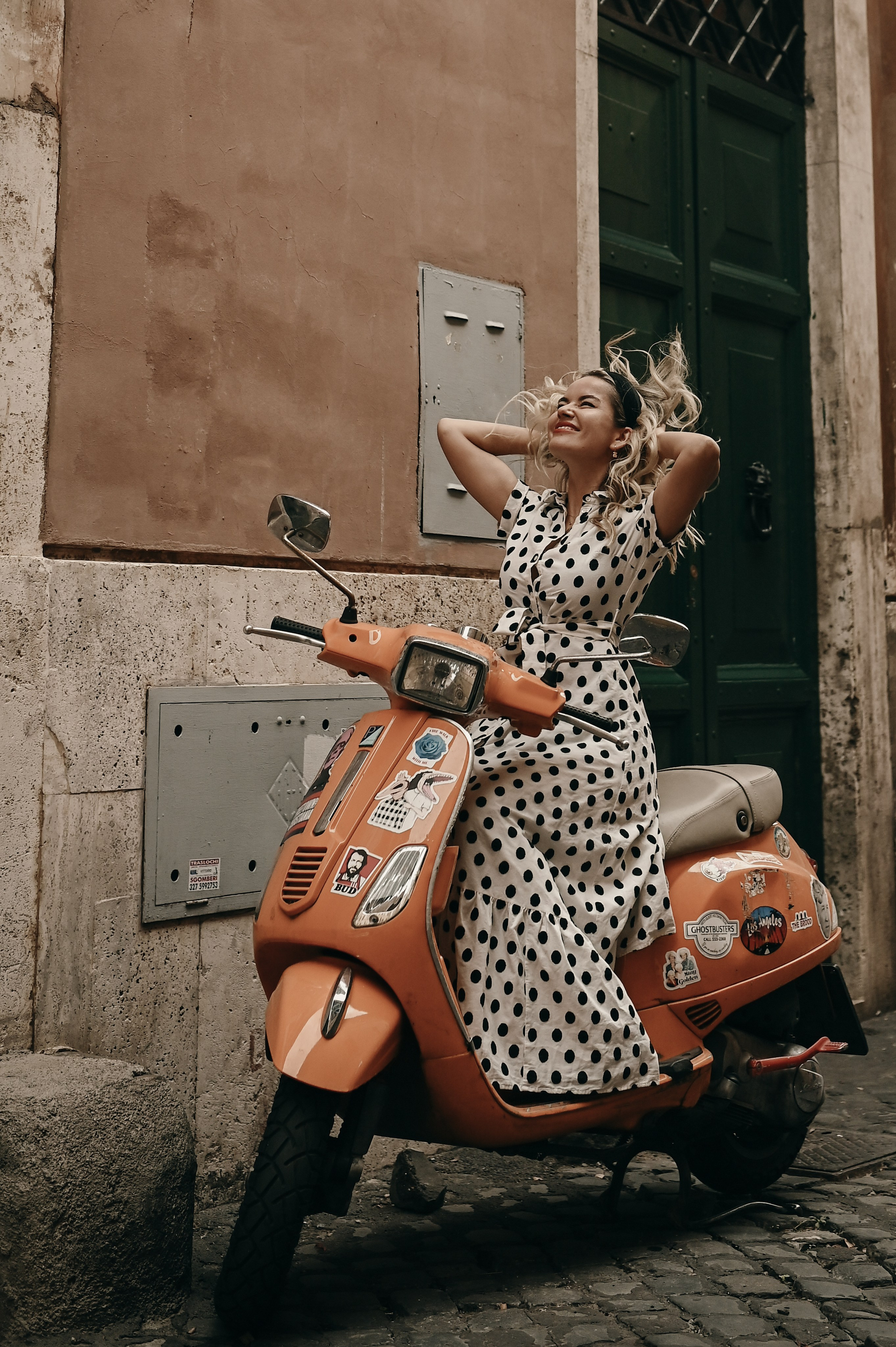 A joyful woman sits on a bright orange Vespa on a cobbled street in Trastevere
