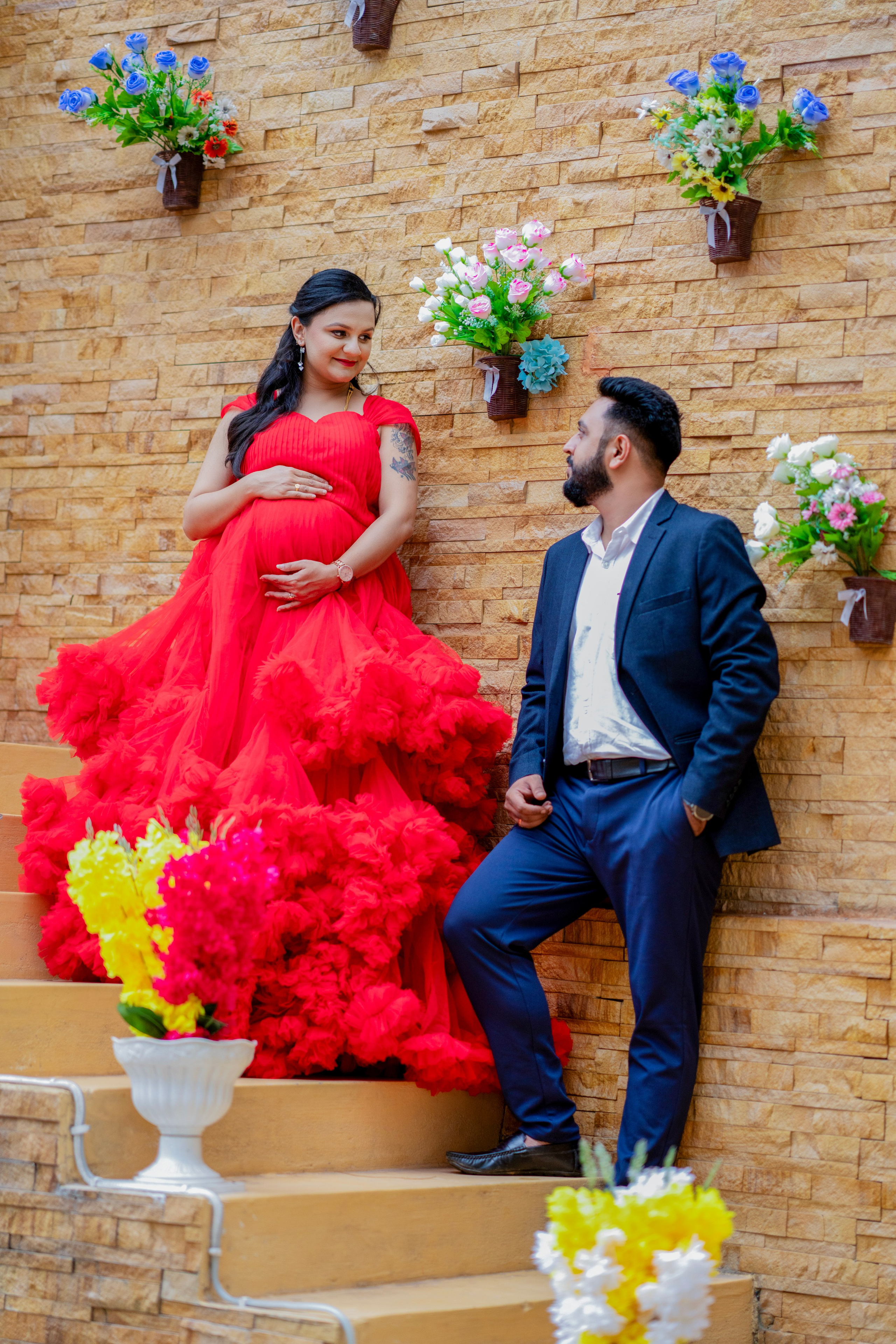 Maternity photoshoot in Bengaluru featuring a woman in a voluminous red ruffled gown and a man in a navy suit posing on stairs against a stone wall with flowers.