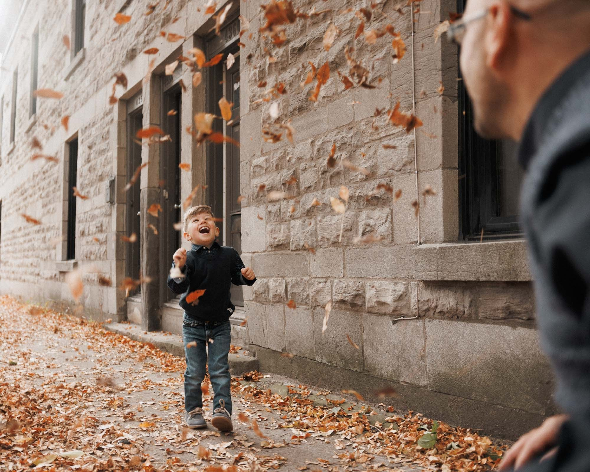 Young boy playing in falling autumn leaves beside a stone wall
