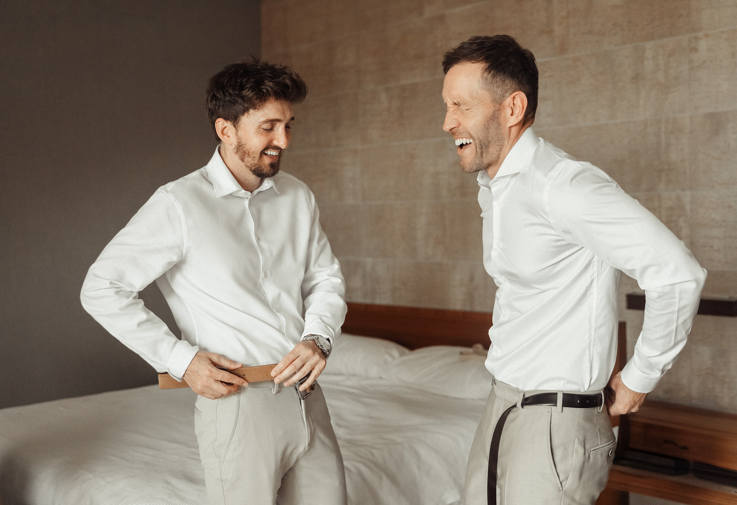 The groom preparing for his wedding day, adjusting his belt, with close friend in a relaxed and joyful atmosphere. Rhodes, Greece