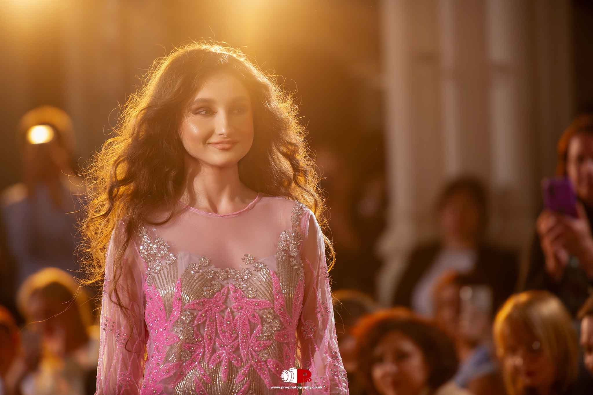 A model in a pink and silver gown is walking down the runway under golden lighting.