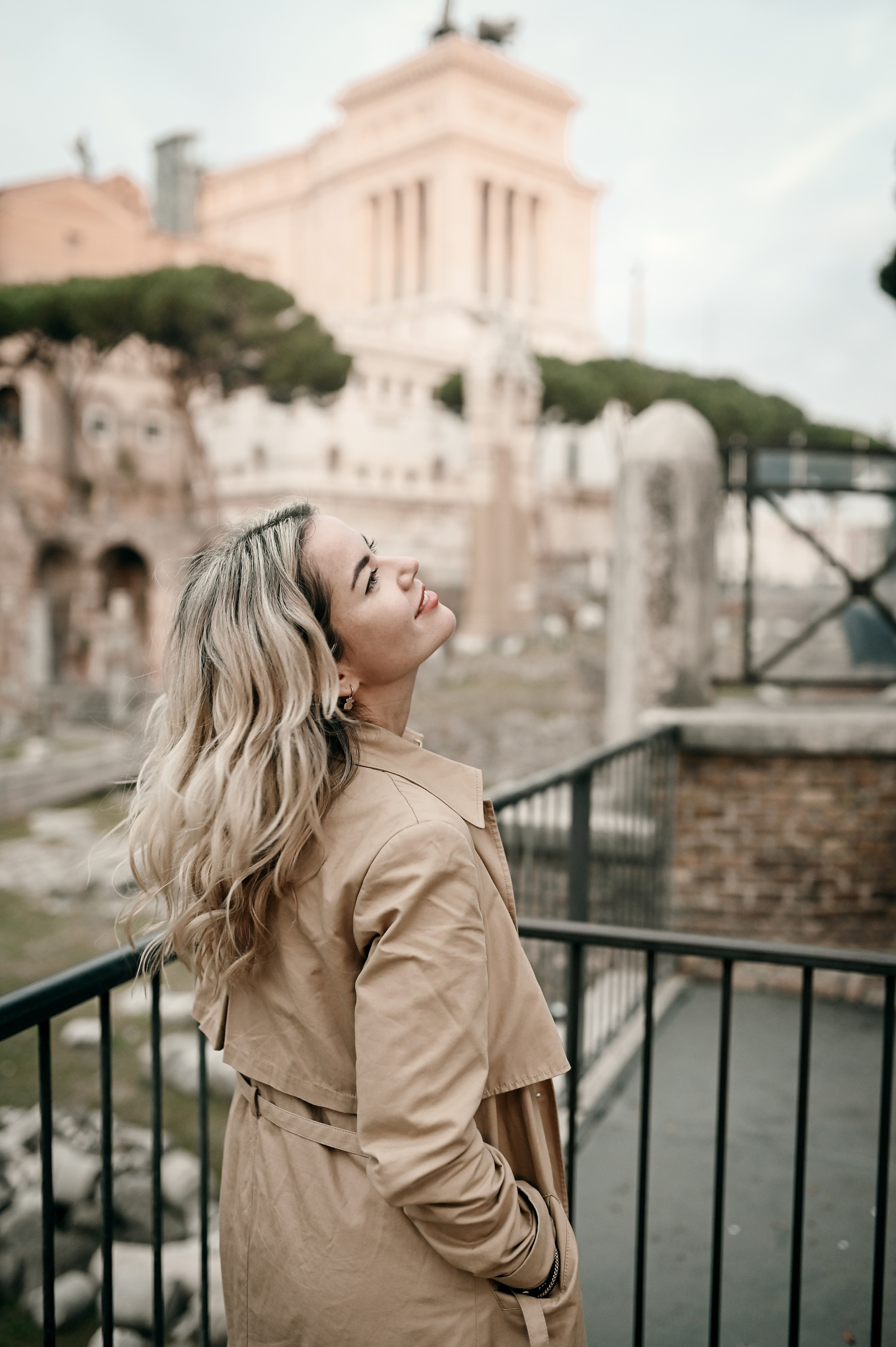 Woman in a beige trench coat gazes dreamily upward, with the Roman Forum softly lit in the morning background