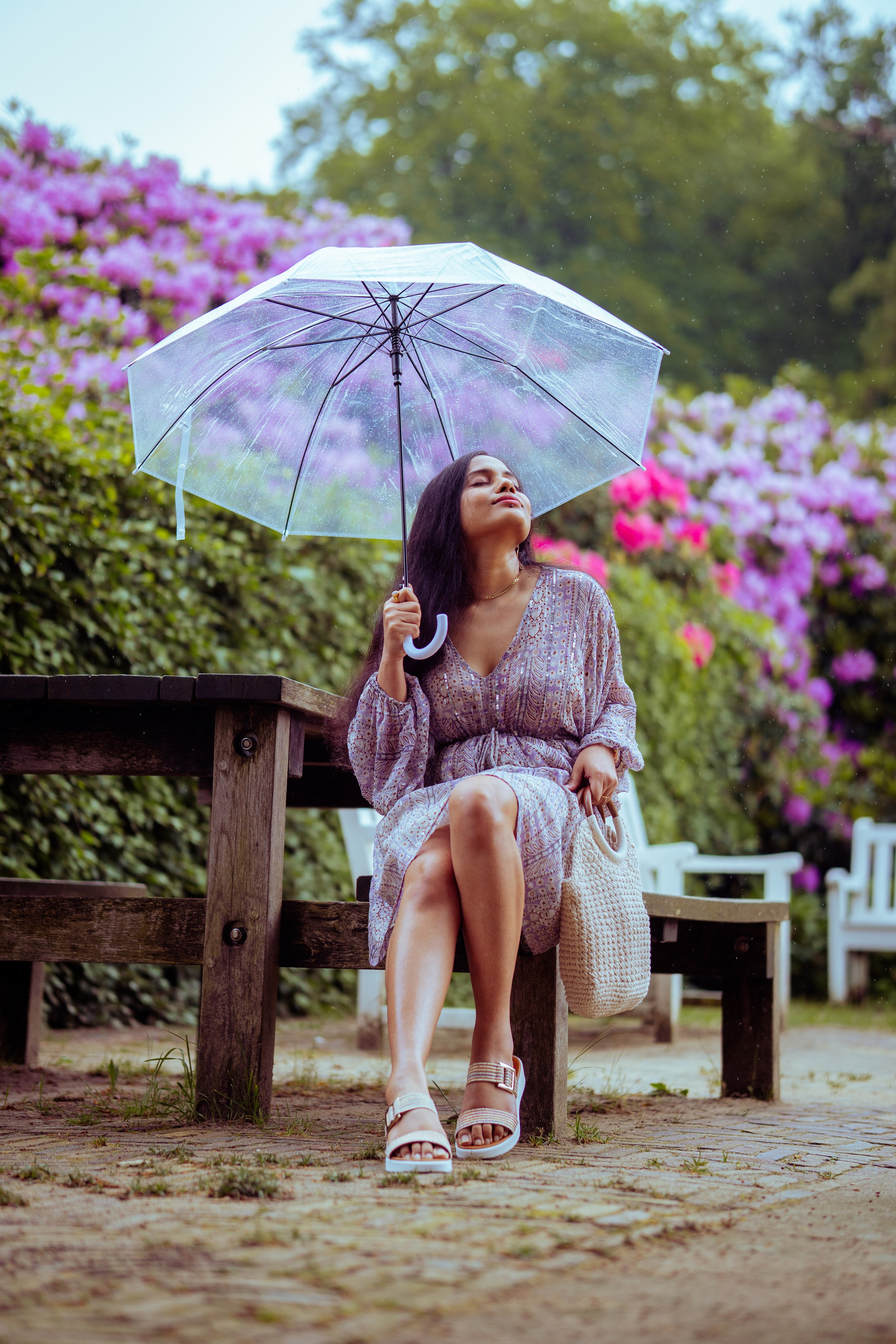 woman sitting on a wooden bench with flowers as background and holding a transparent umbrella