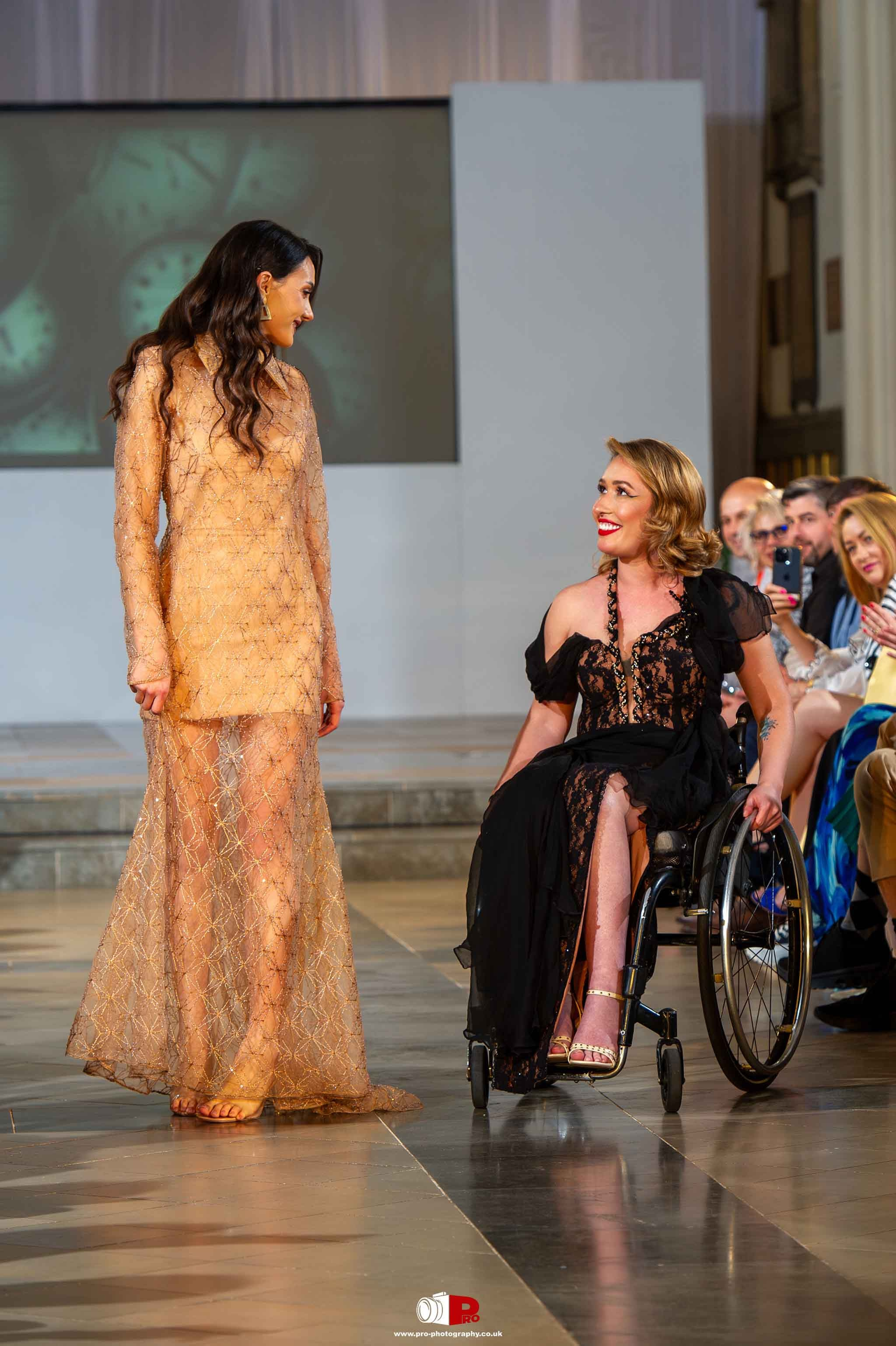 A female model in a black lace gown is seated in a wheelchair, smiling at another model in a golden sheer gown on the runway.