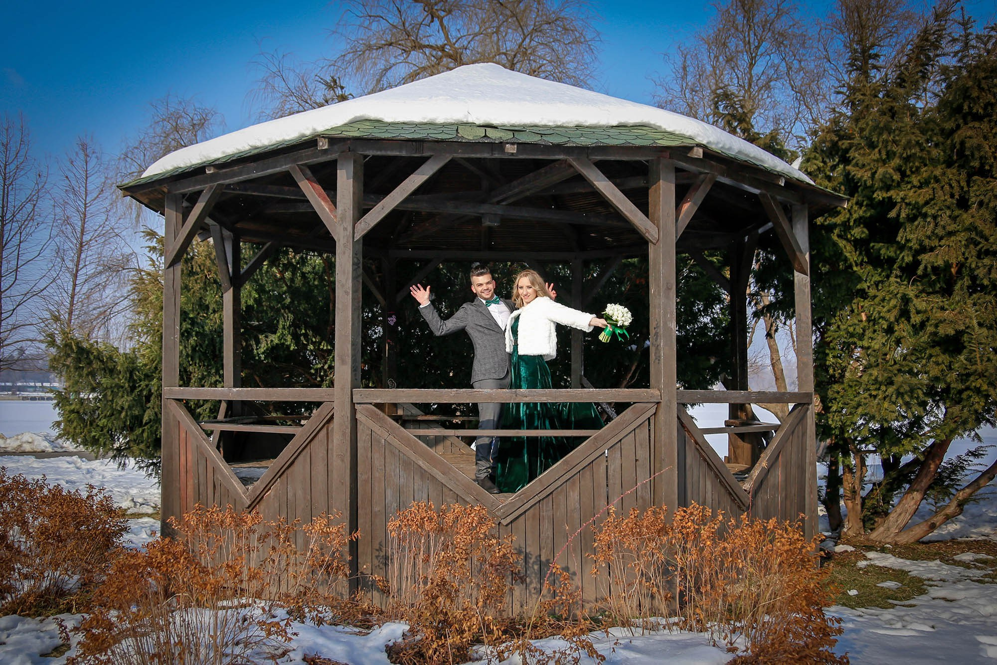 Joyful shot of the newlyweds with their hands raised in celebration, posing in front of the rustic wooden gazebo in winter.