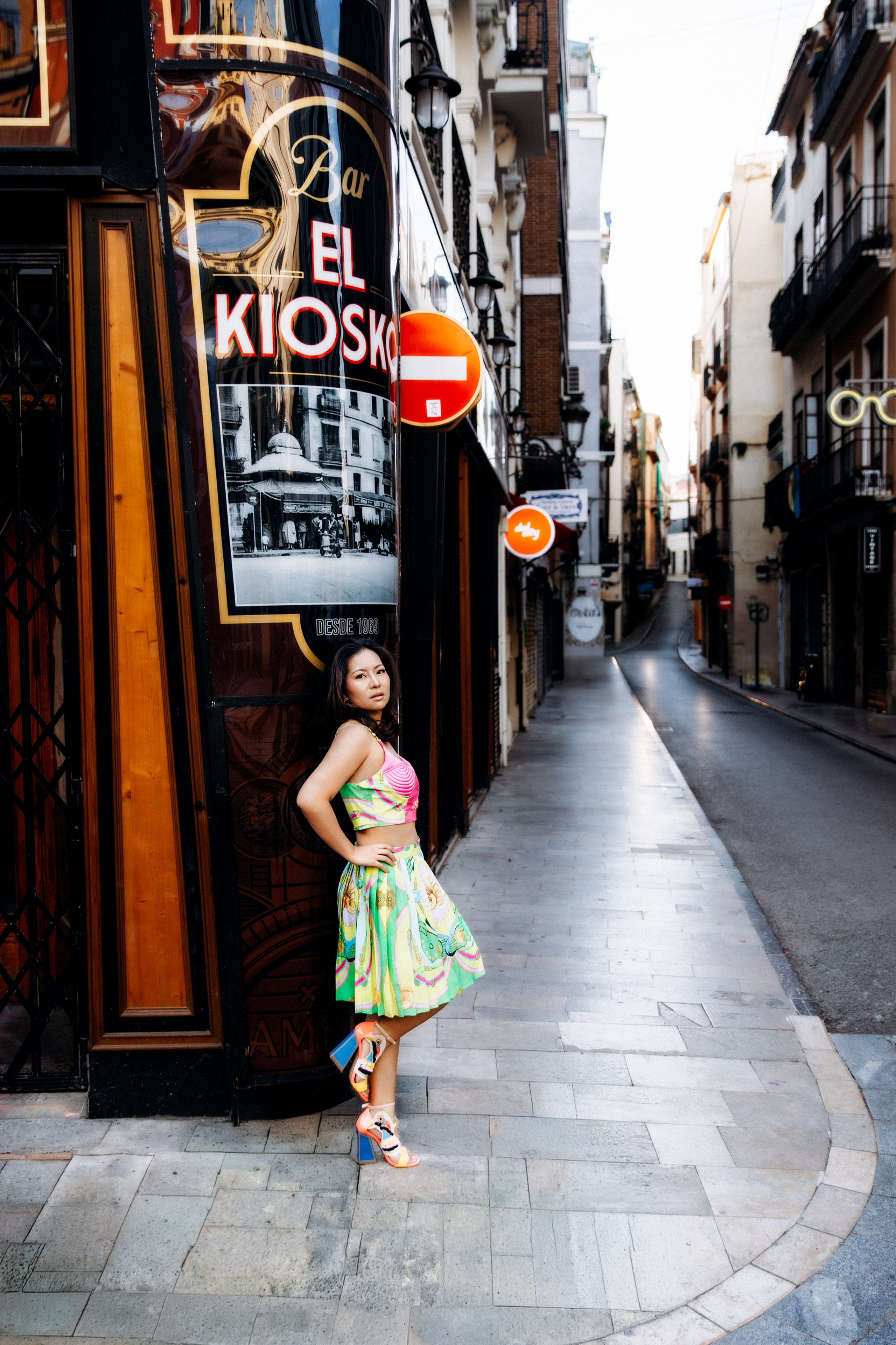 Sesión de retrato femenino en Valencia, España, con una mujer vestida con un ligero vestido de verano posando con confianza en una encantadora calle histórica rodeada de arquitectura mediterránea, comercios locales y detalles urbanos. Capturada con luz natural, esta vibrante imagen lifestyle destaca la individualidad, la confianza y la belleza inspirada en los viajes, ideal para fotografía de retrato femenino, sesiones individuales, marca personal y retratos vacacionales en Valencia y toda España.