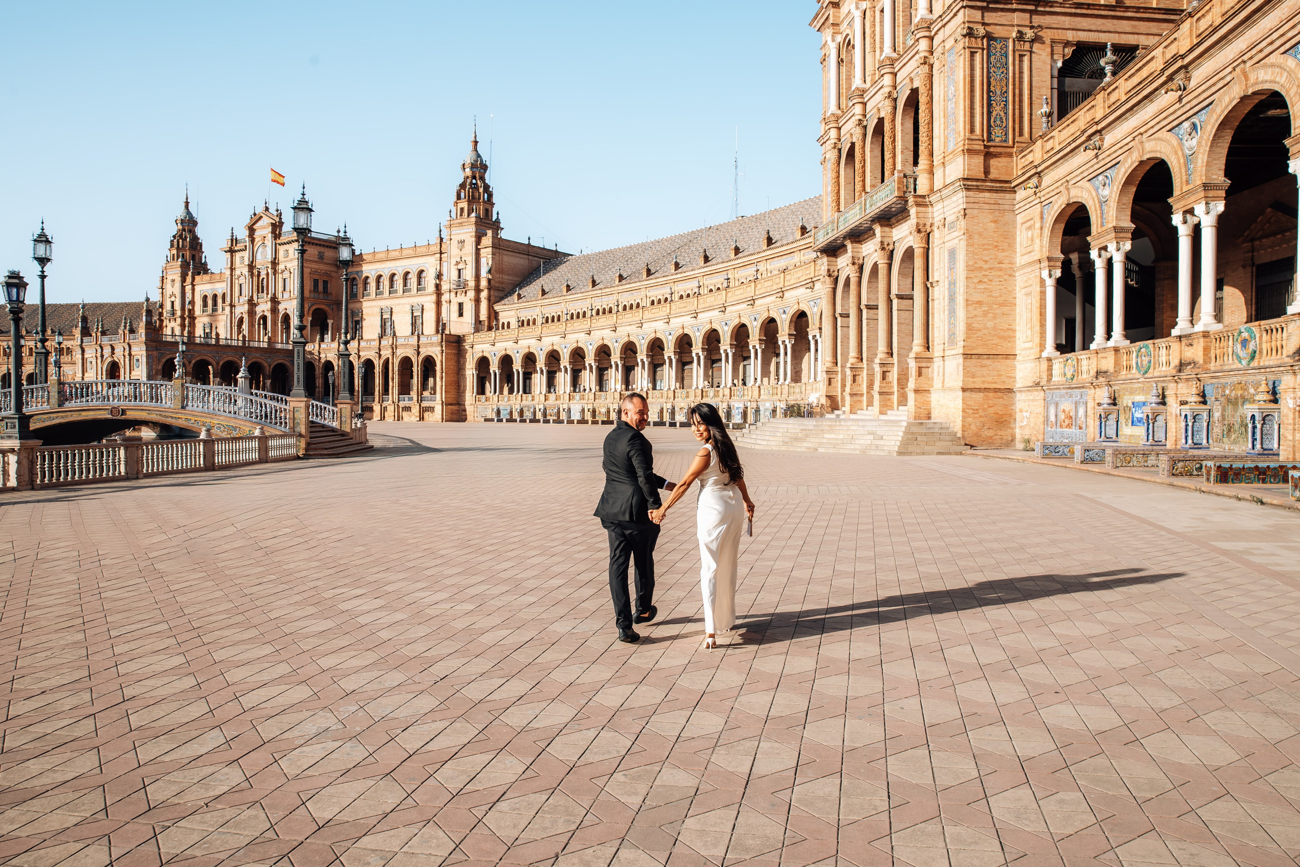 Bride and groom walking hand in hand across the grand plaza in Sevilla with historic architecture in the background. Natural documentary-style moment from their intimate destination wedding in Spain.