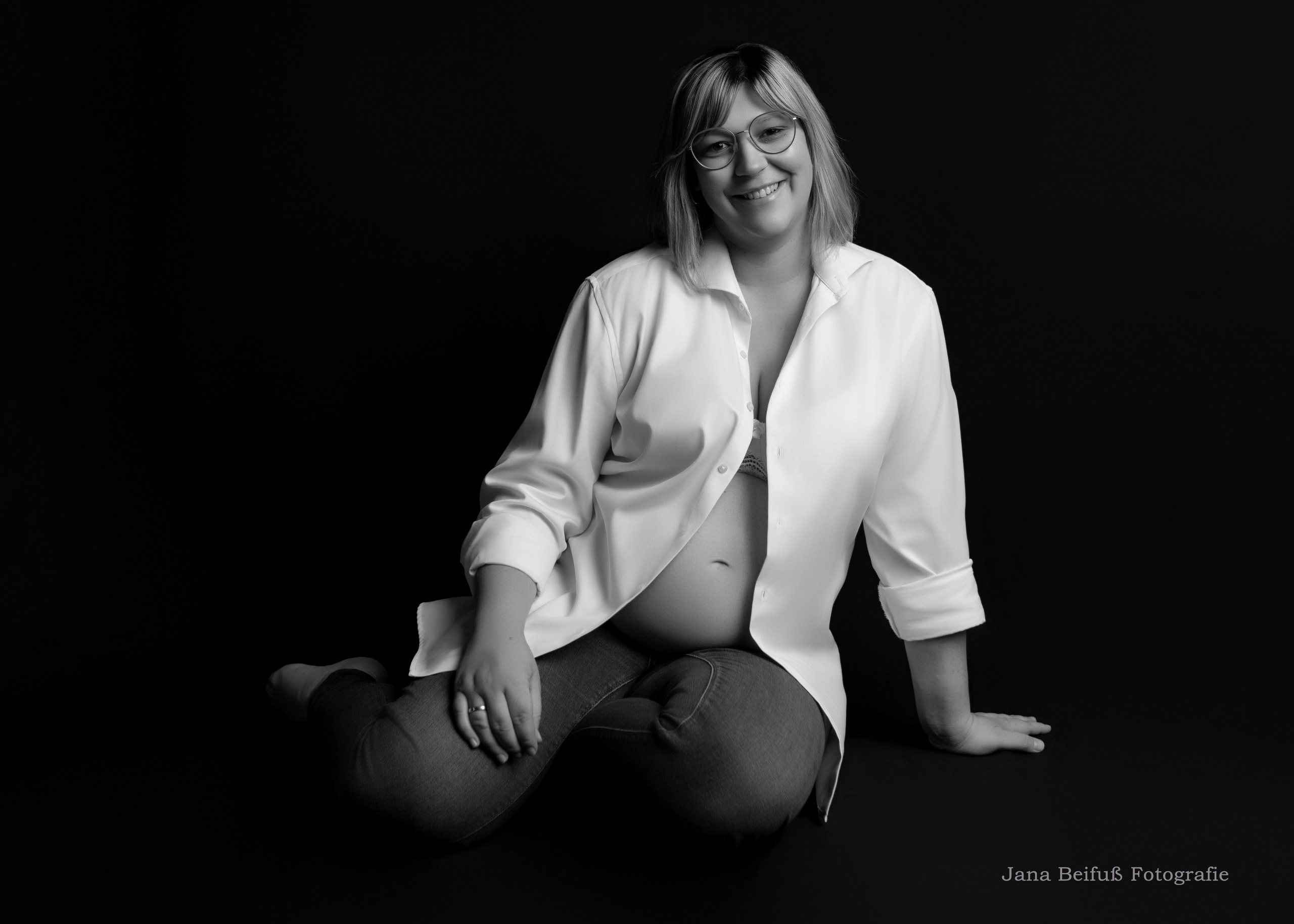 Werdende Mama sitzt auf dem Boden und lächelt schwarzer Hintergrund Fotografin Jana Beifuß in Weißenhorn bei Memmingen Illertissen Ulm