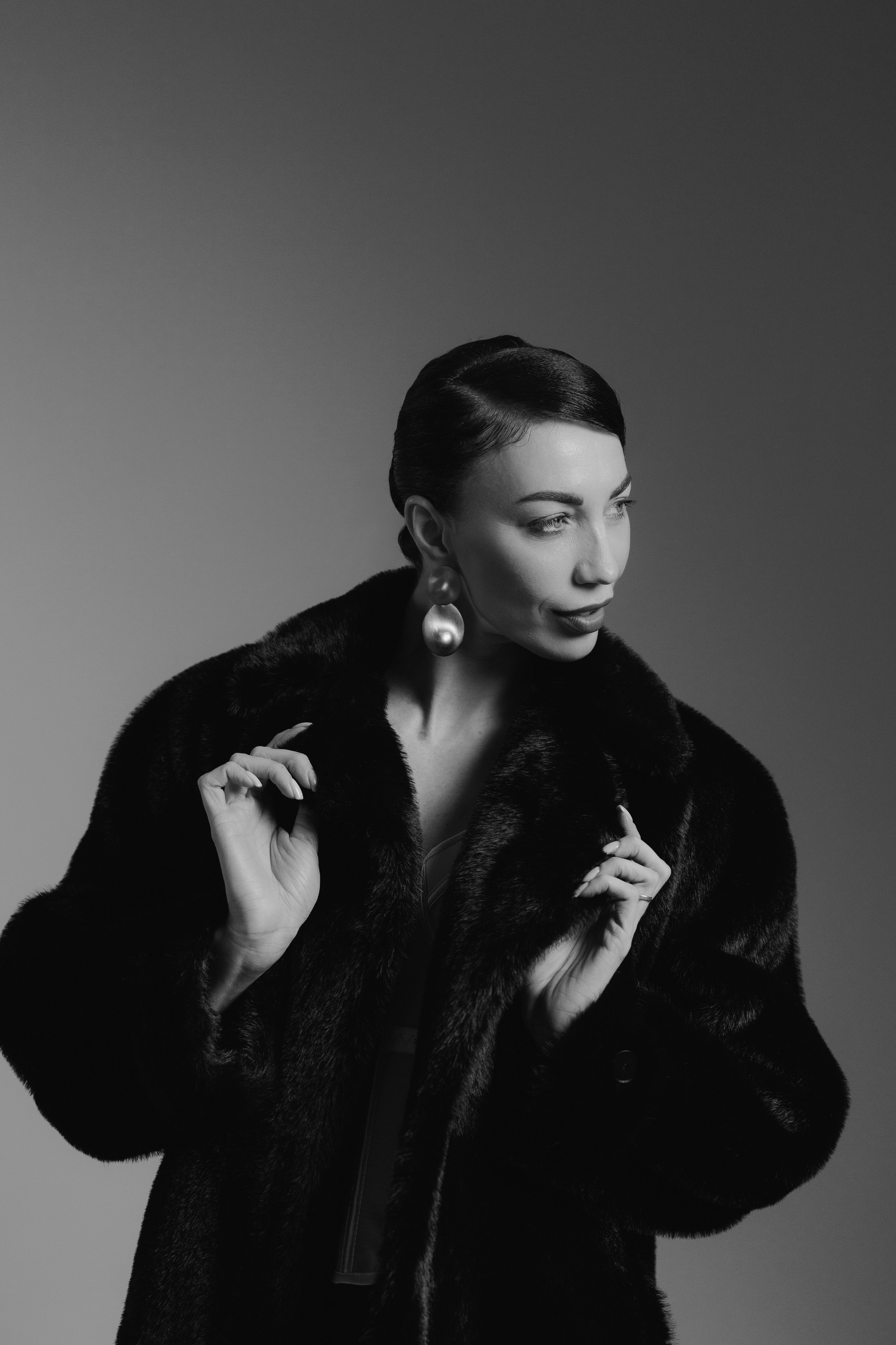 Fashion studio portrait of a woman in a black fur coat adjusting the collar with dramatic shadows and soft studio light, photographed in Leipzig, Germany. Фэшн-студийный портрет девушки в чёрной шубе, поправляющей воротник, с драматичными тенями и мягким студийным светом, снятый в Лейпциге, Германия.