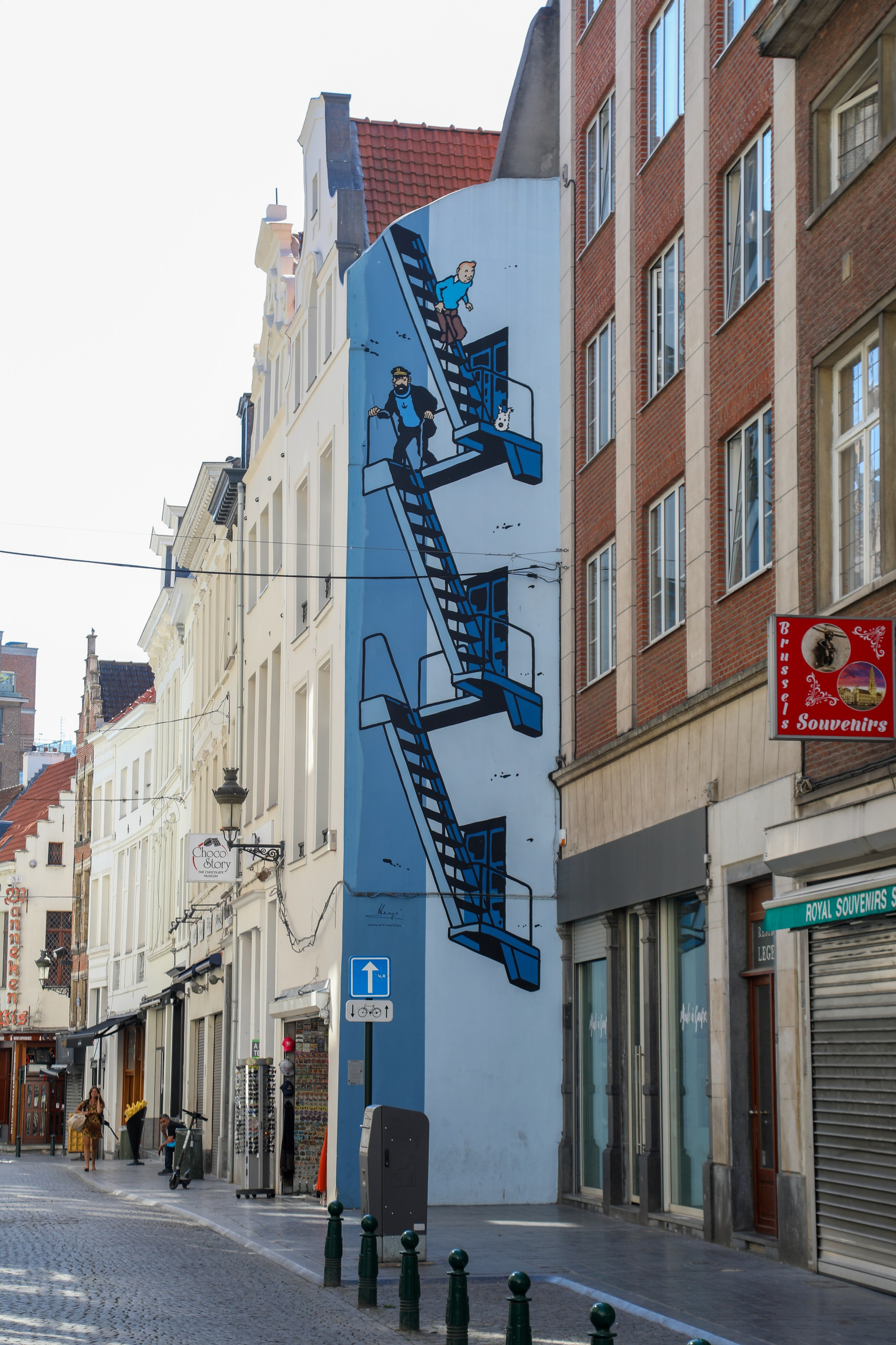 A large, blue-and-white comic strip mural painted on the side of a building in Brussels, depicting Tintin, Captain Haddock, and Snowy the dog climbing an exterior fire escape.