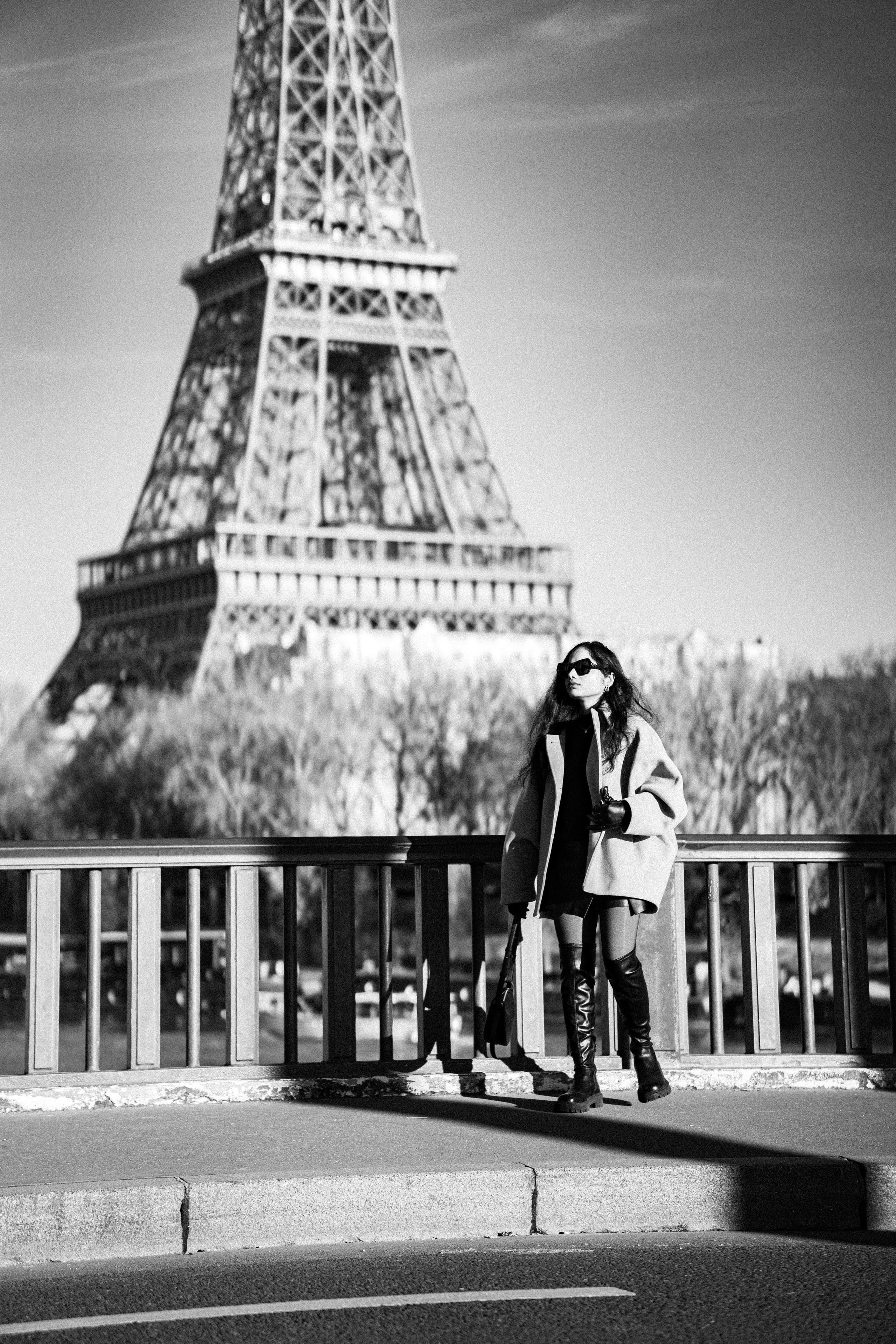 a woman wearing oversized winter jacket and walking on a bridge with eiffel tower view behind her