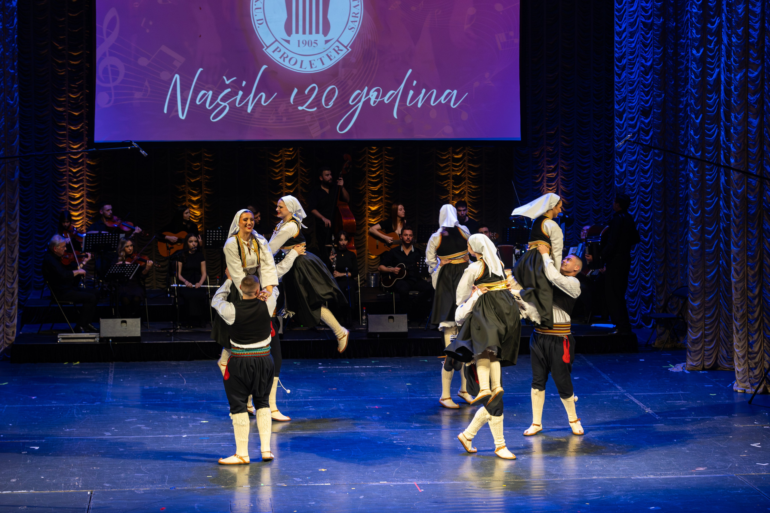 Galerija fotografija sa svečanog koncerta RKUD Proleter u Narodnom pozorištu Sarajevo, prikazuje nastupe izvođača, svečanu atmosferu, bogatu scenografiju i publiku u prepunoj dvorani.