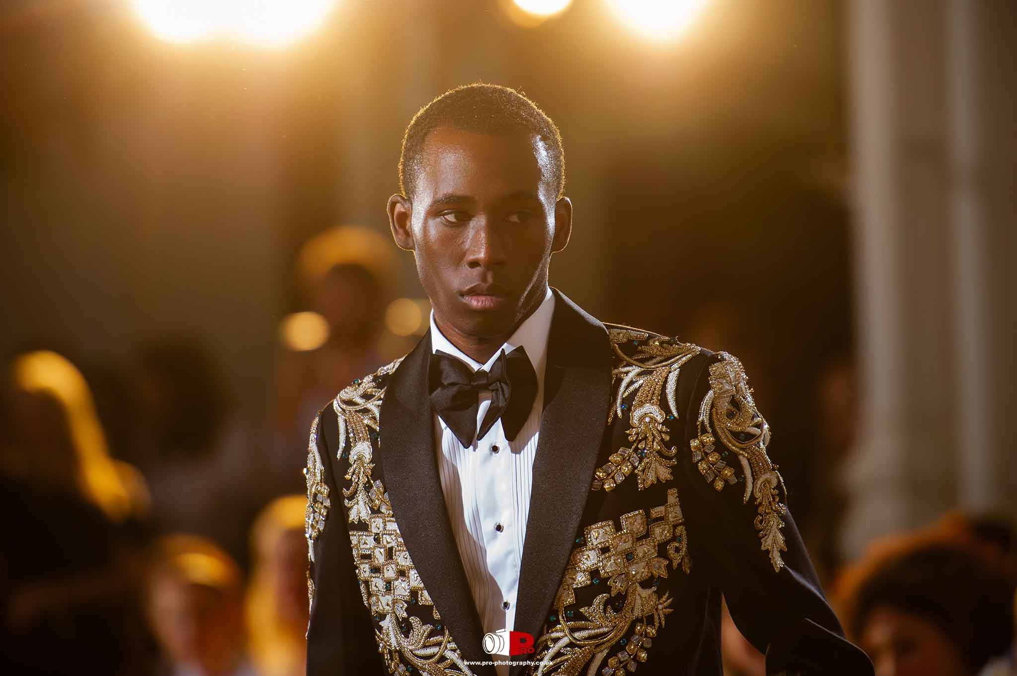 A male fashion model confidently walks down the runway in a black tuxedo adorned with intricate gold embroidery.