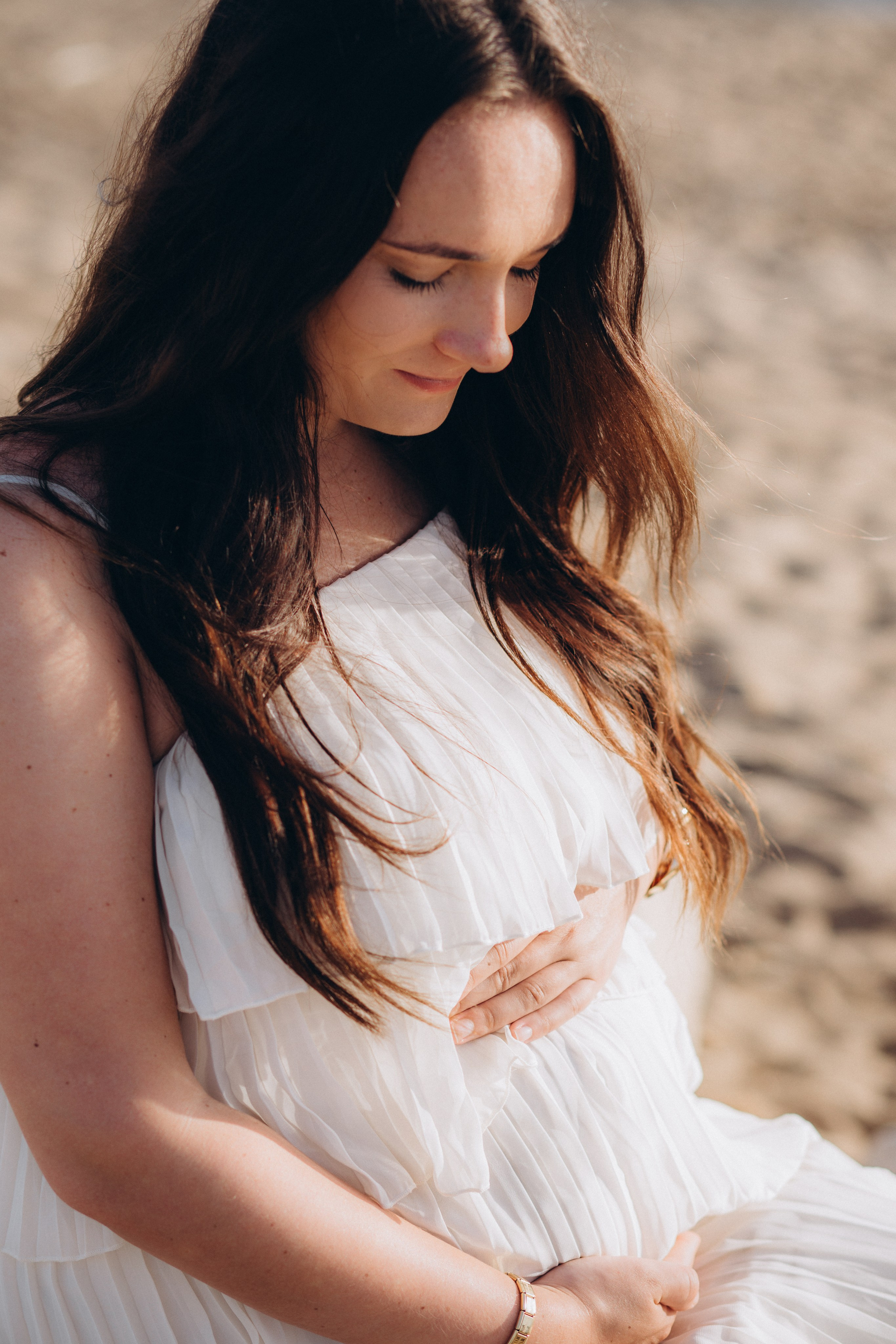 Retrato tranquilo de embarazo en la playa de Barcelona, España — primer plano de una mujer embarazada con vestido blanco sosteniendo suavemente su vientre. Ideal para quienes buscan sesiones suaves y emocionales de maternidad en Barcelona y la costa.