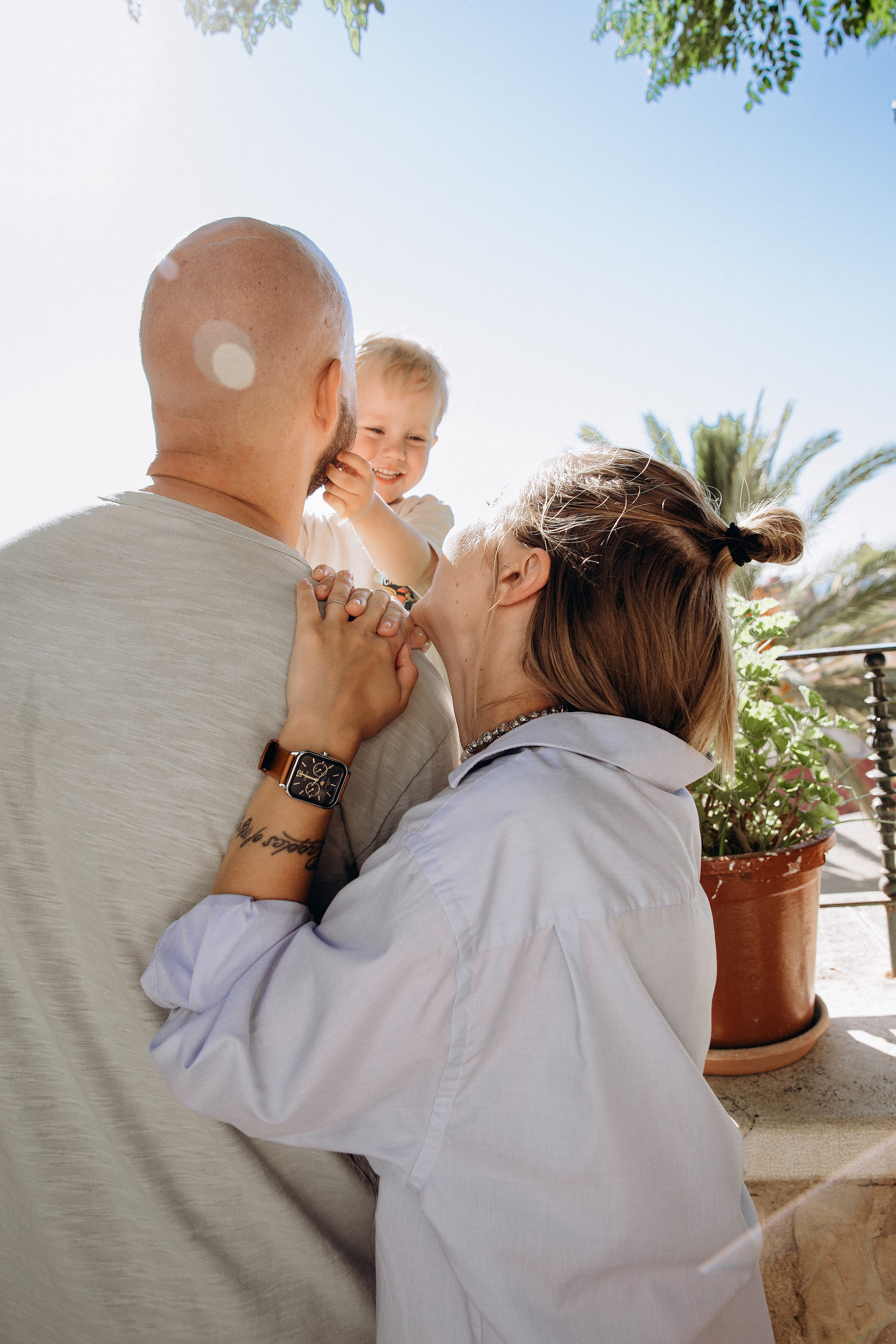 Momento familiar espontáneo durante una sesión soleada en Valencia, España — madre y padre abrazados sonriendo a su hijo alegre. Perfecto para quienes buscan sesiones familiares emocionales y naturales en Valencia y España.