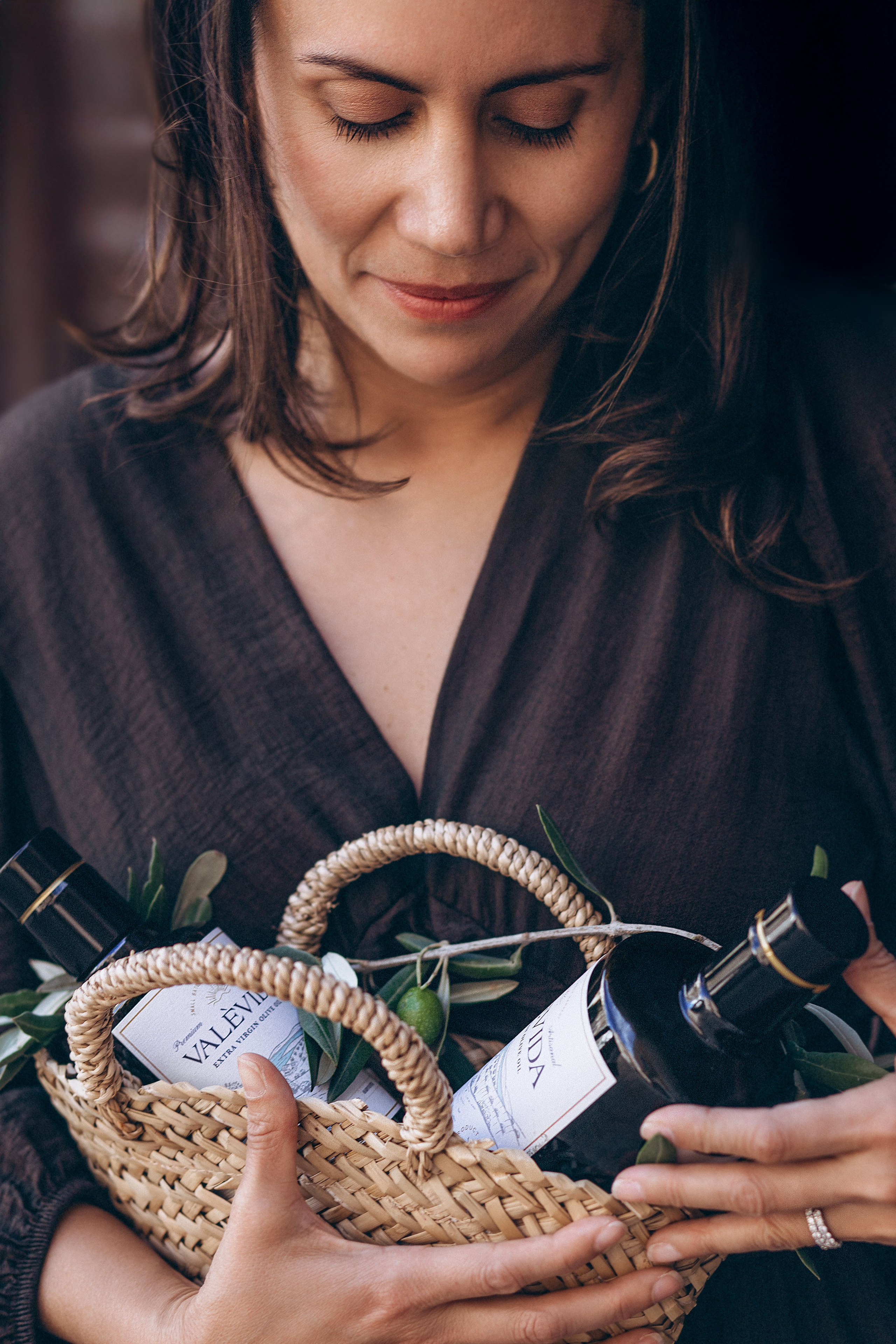Elegant brand photography captured in Valencia, Spain, featuring a woman in a dark dress holding a woven basket filled with premium olive oil bottles and olive branches — ideal for showcasing product lifestyle shoots and content branding for businesses seeking natural, Mediterranean-inspired visuals in Spain.