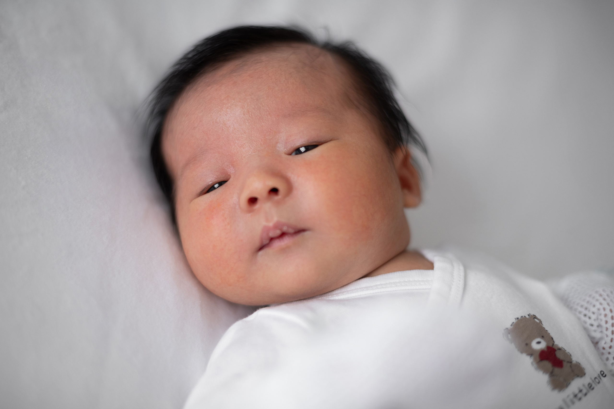 Close-up of newborn baby boy lying on white bedding, looking sleepily at the camera in soft window light.