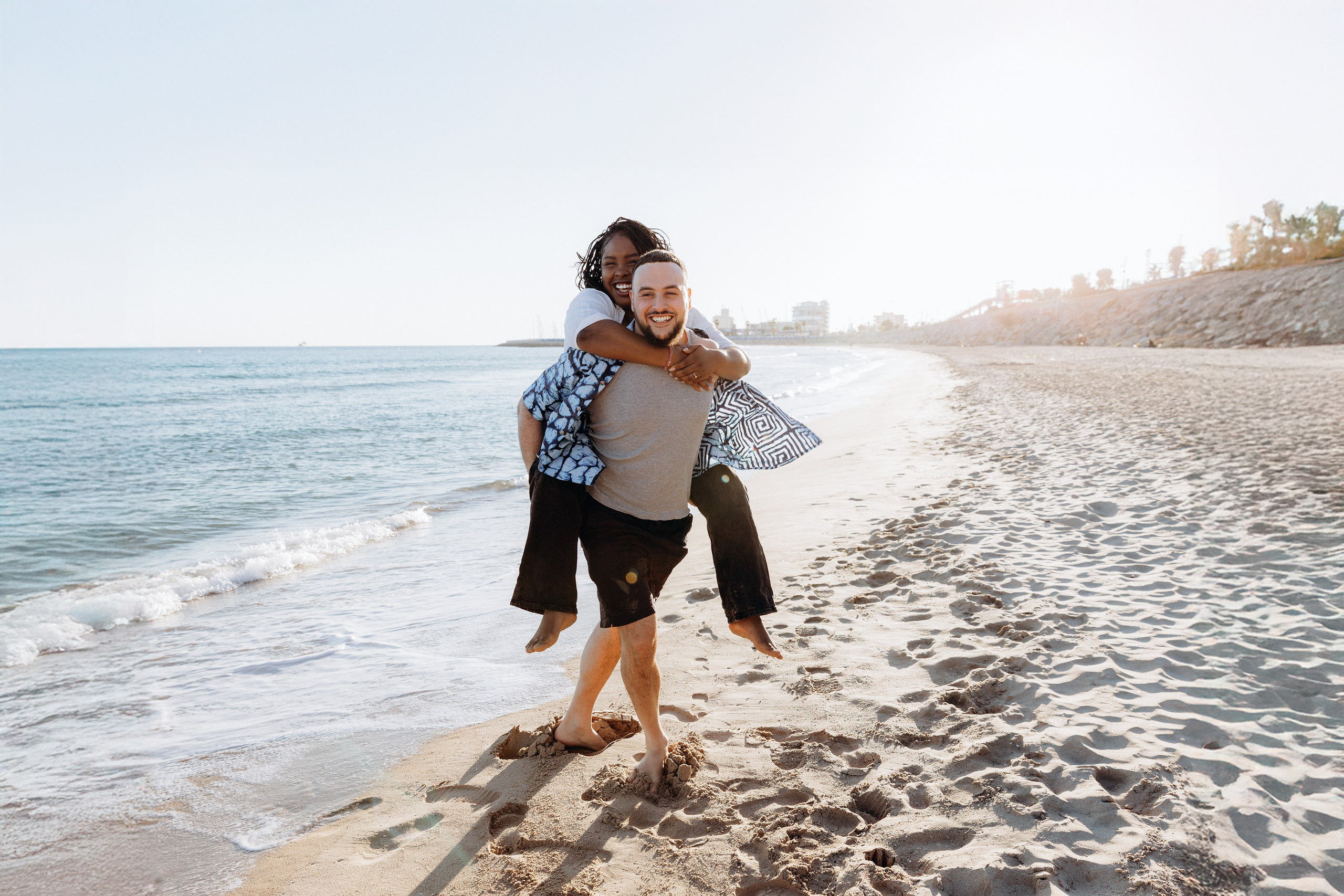 Sesión de fotos alegre en la playa de Valencia, España — un hombre lleva a su pareja a caballito por la orilla, capturando risas y amor. Ideal para parejas que buscan sesiones divertidas y espontáneas de historia de amor en Valencia y en toda España.