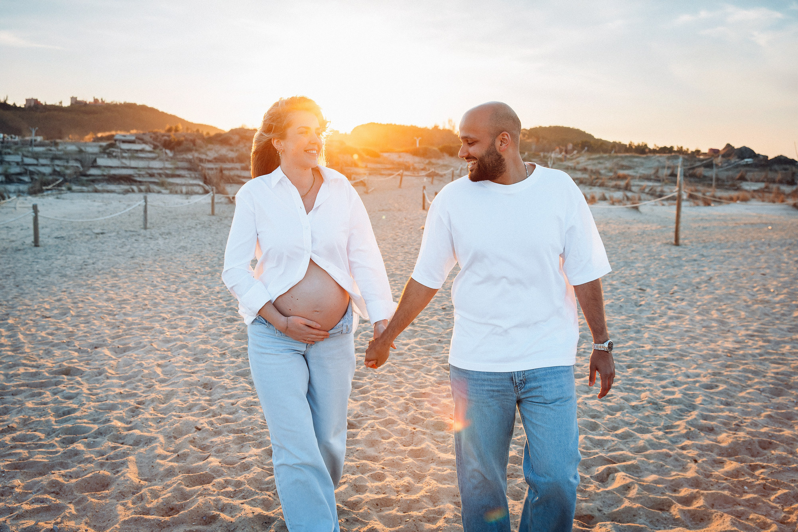 Sesión de fotos de embarazo en Valencia, España, con una pareja caminando de la mano por la playa al atardecer, con la mujer embarazada sosteniendo delicadamente su barriga, capturada con cálida luz dorada — ideal para sesiones maternity en Valencia y España.