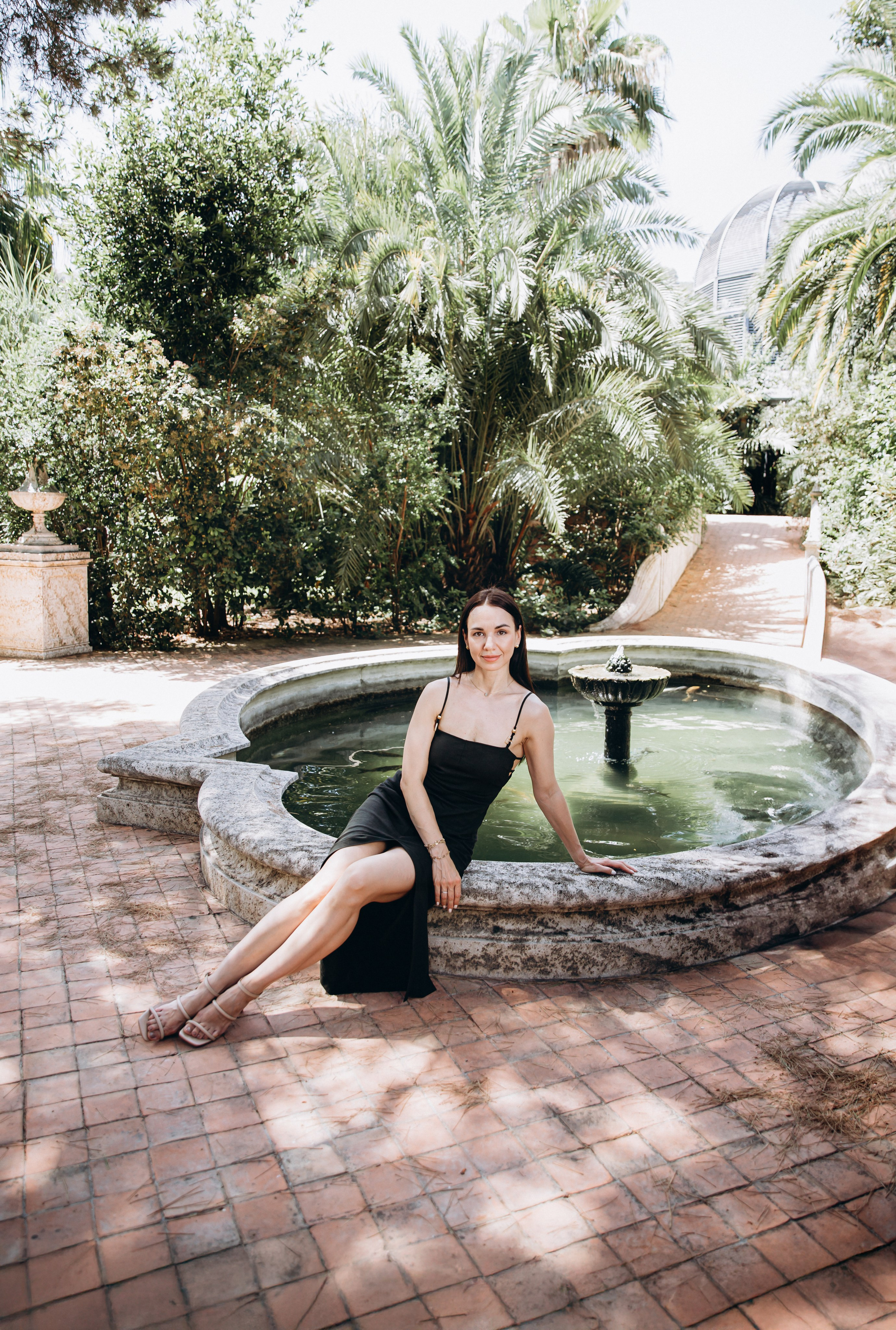 Элегантная уличная портретная фотосессия в Мадриде, Испания — женщина в чёрном платье сидит у исторического фонтана, окружённого пышными пальмами, идеально для тех, кто ищет стильные садовые портреты в Мадриде и по всей Испании.