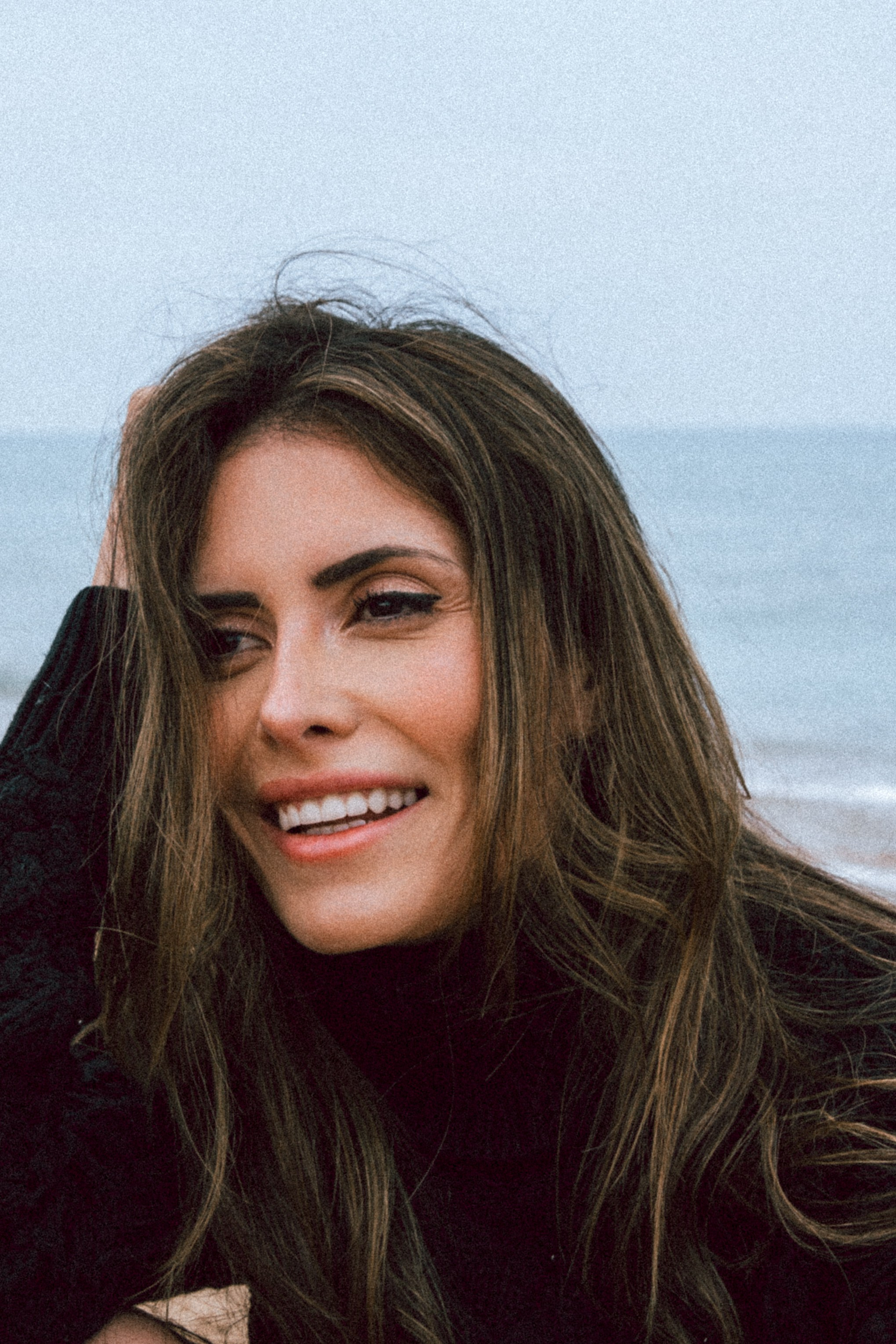 Непринуждённый пляжный портрет улыбающейся женщины в чёрном свитере с растрёпанными волосами и расслабленным выражением лица, с морем на заднем плане