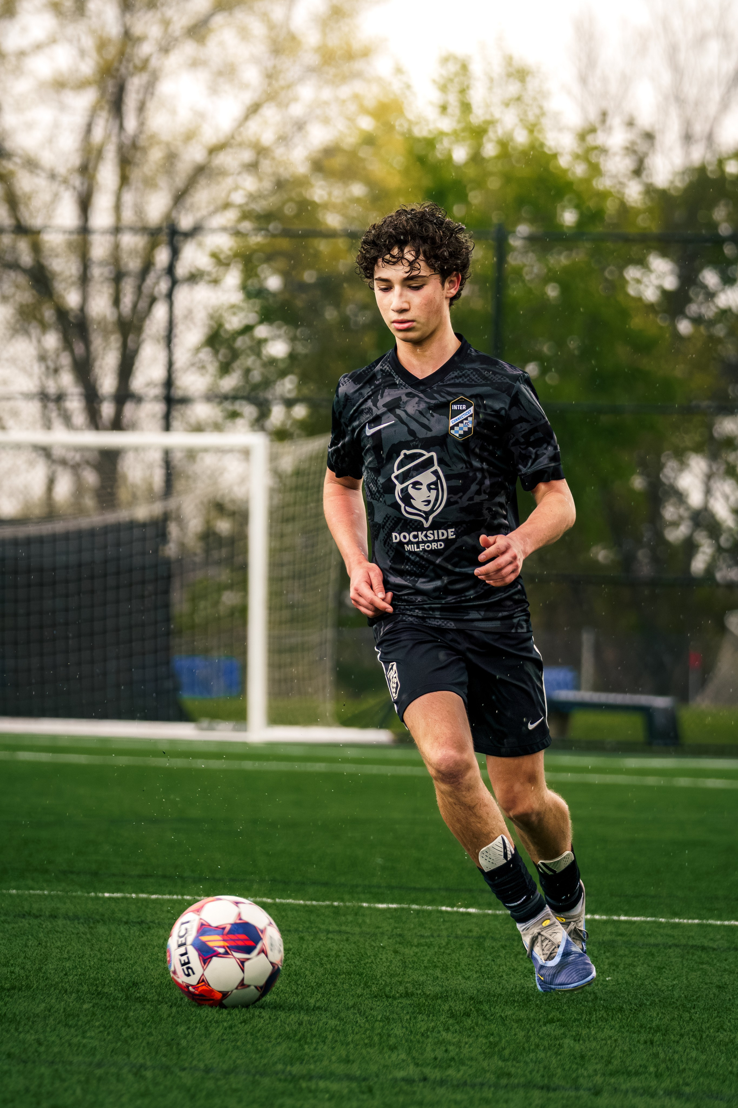 High school soccer player in a black kit dribbling forward during an outdoor match.
