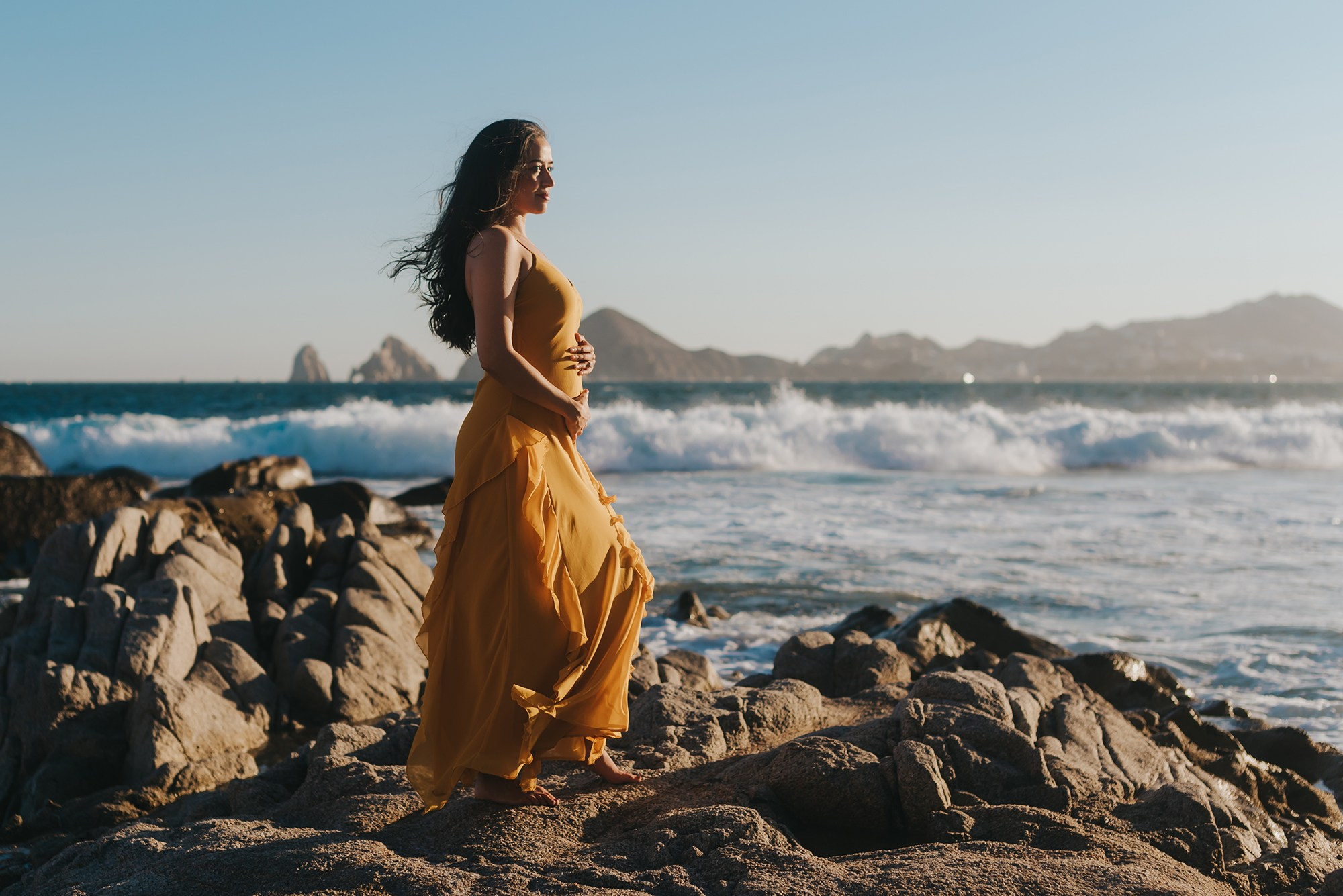 Pregnant woman embracing her belly at sunset with Cabo San Lucas Arch and ocean waves