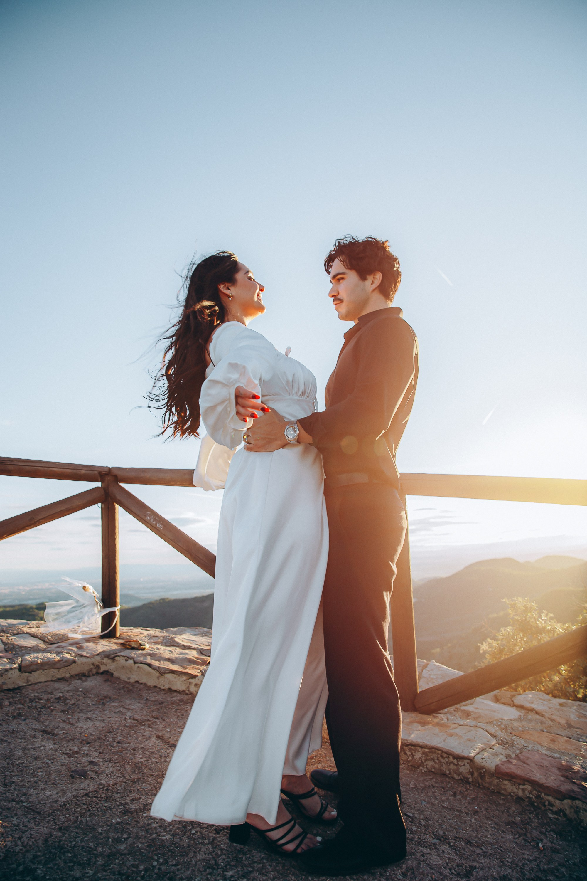 Engagement photoshoot in València, Spain, capturing a romantic couple standing close at a scenic viewpoint during golden hour, with soft sunset light, a flowing white dress, and natural mountain views in the background — an intimate wedding love story image ideal for engagement photography, pre-wedding sessions, elopements, and professional wedding photoshoots in València and across Spain.