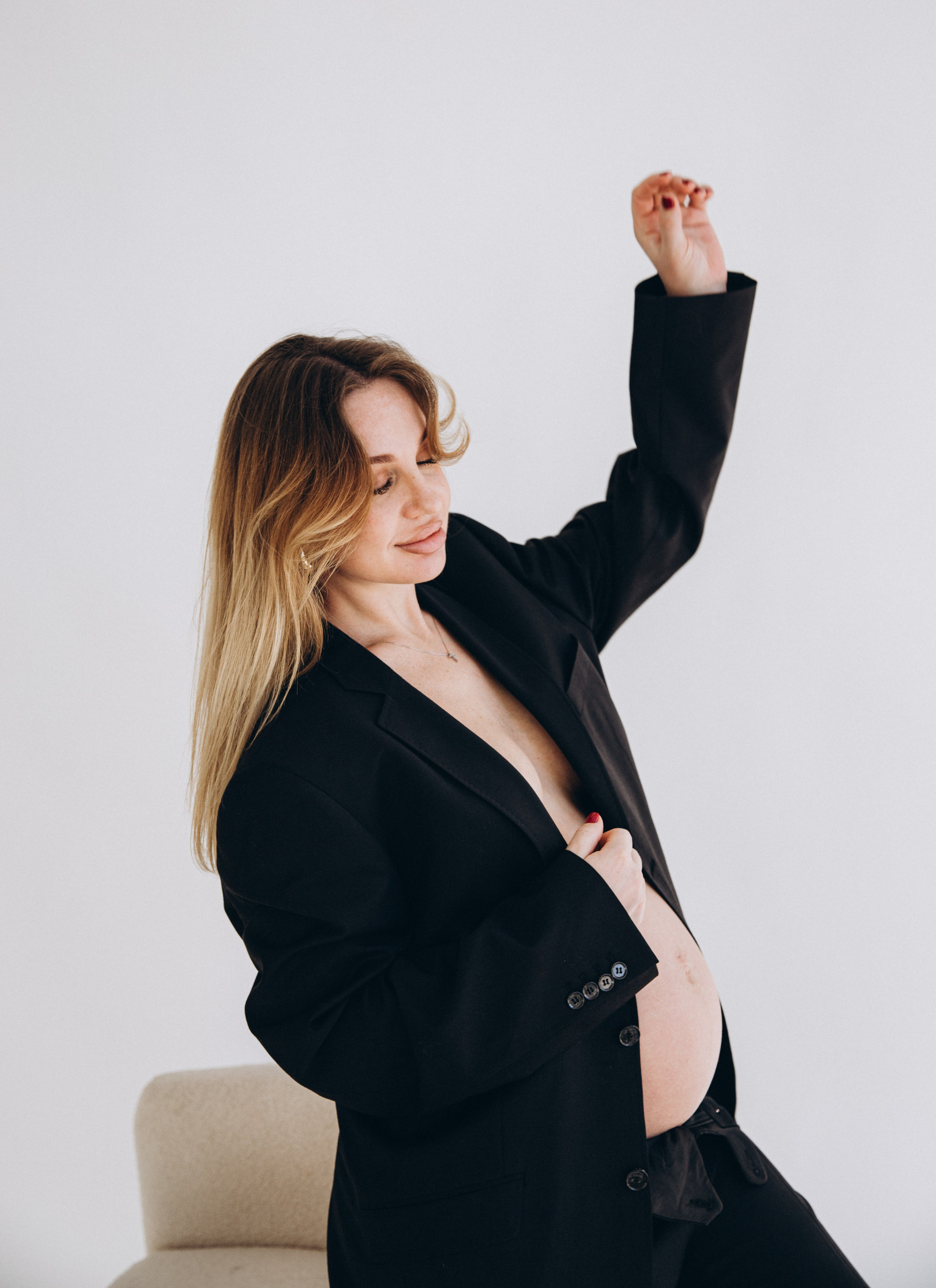 Retrato de embarazo con confianza en Valencia, España — mujer embarazada con americana negra oversize y una sonrisa suave, posando ante un fondo minimalista de estudio.