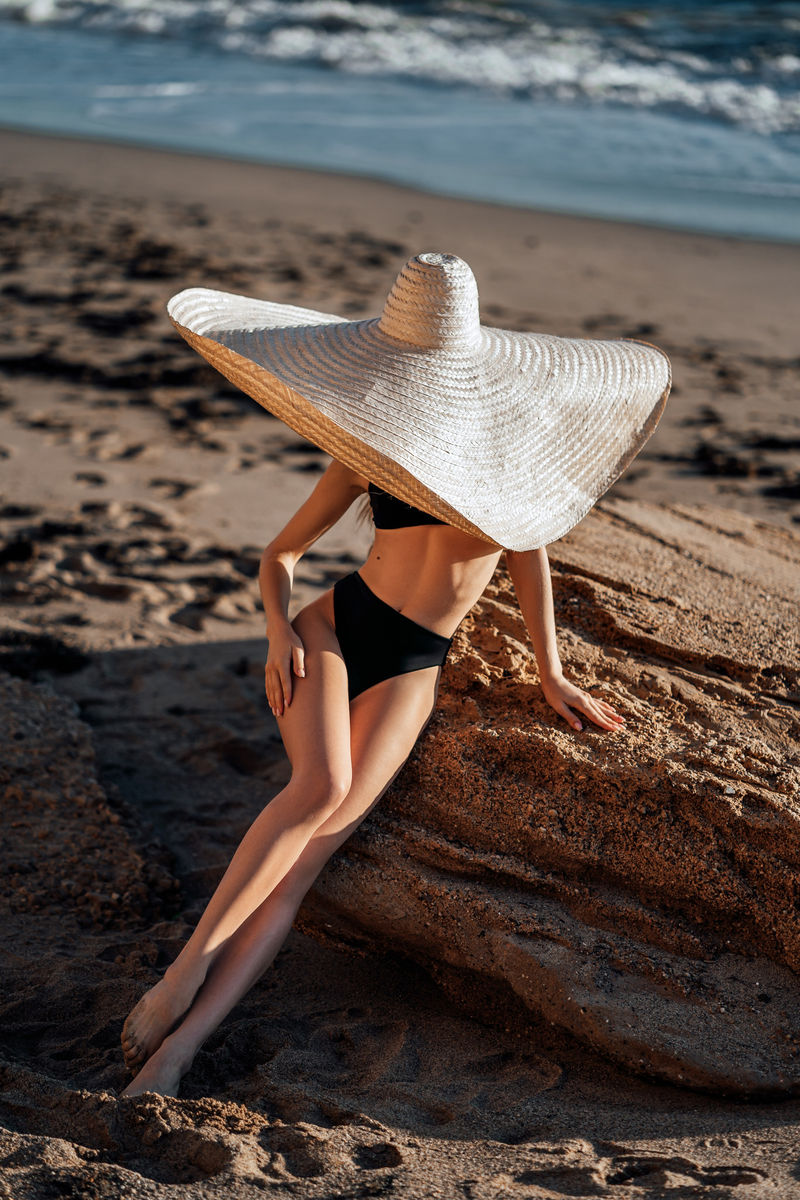 Making a stylish statement by the sea, the model’s black bikini and wide-brimmed straw hat blend perfectly with the oceanic landscape. Her poised stance adds to the allure of this beachside shot. Seaside style, black bikini, straw hat, oceanic landscape, poised stance, beachside allure