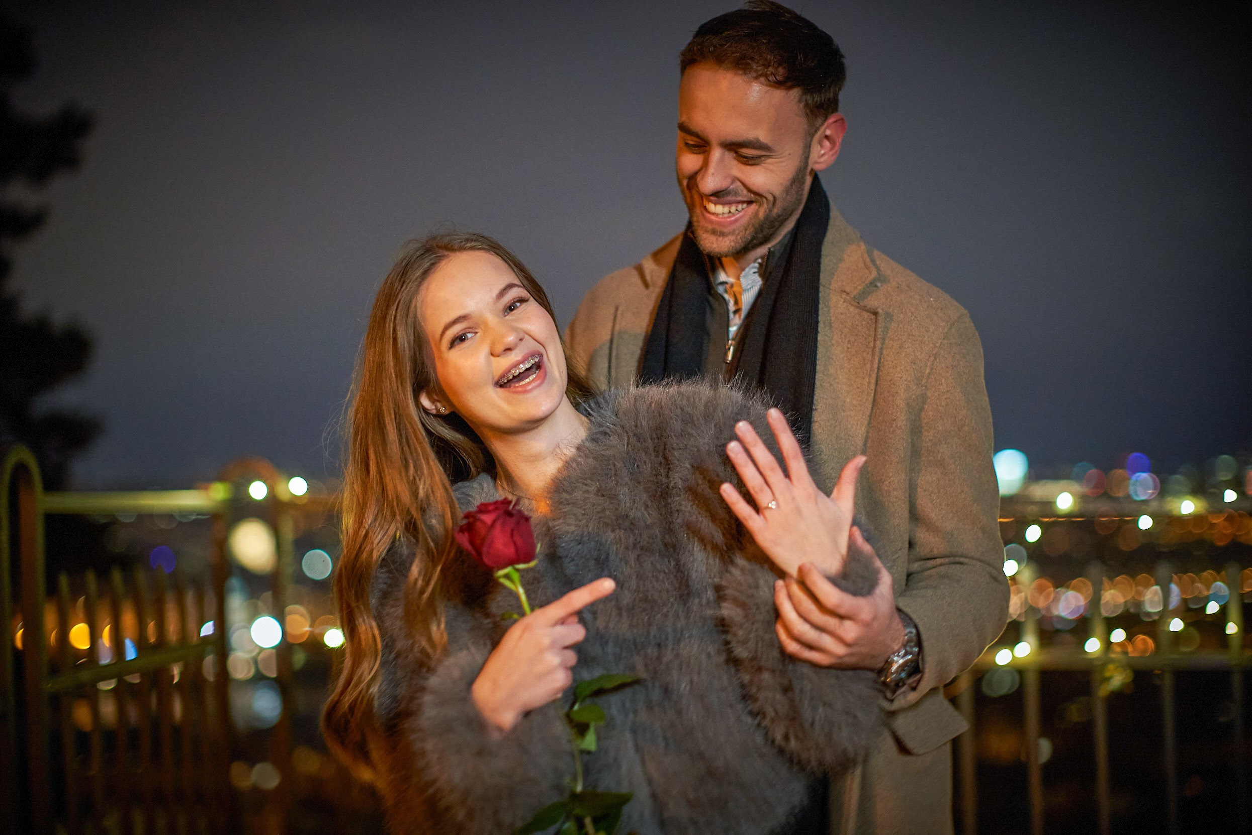 Woman with braces holding rose points to engagement ring as man smiles against Prague skyline.