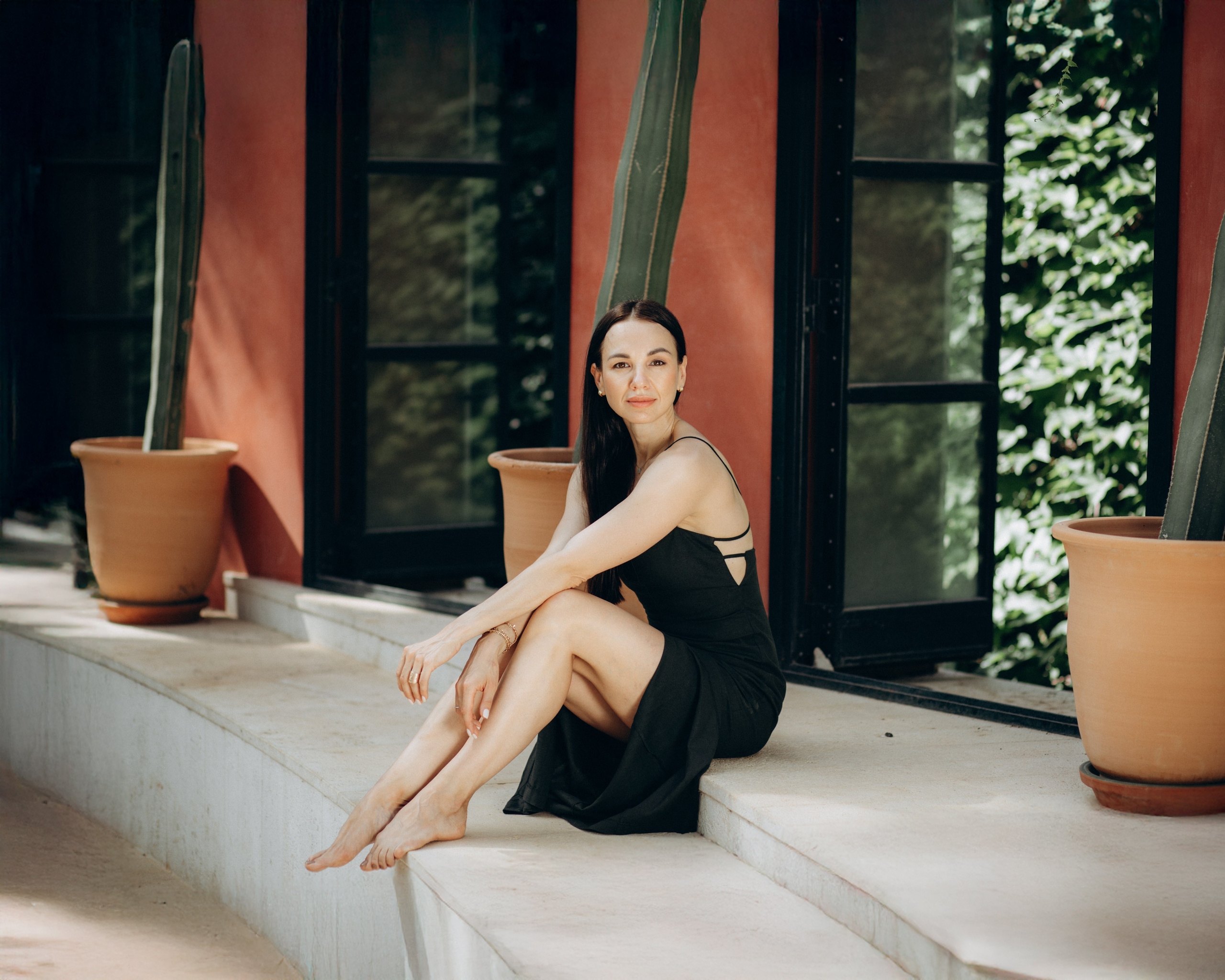 Женщина в черном платье сидит босиком на мраморной скамье перед терракотовой стеной и кактусом в Барселоне, Испания — расслабленная и элегантная портретная фотография, передающая средиземноморский стиль, природную красоту и атмосферу спокойствия, идеально подходит для портретных фотосессий в Барселоне и Испании.