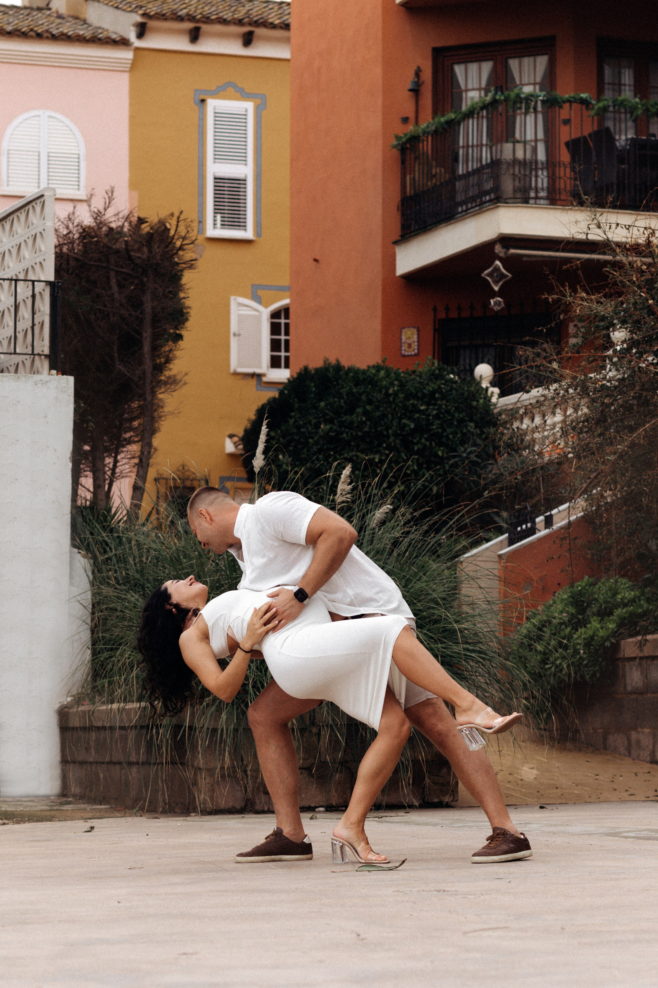 Playful romantic moment of a couple dancing on a charming street with colorful Mediterranean-style houses in the background, captured in Port Saplaya, Valencia, Spain.