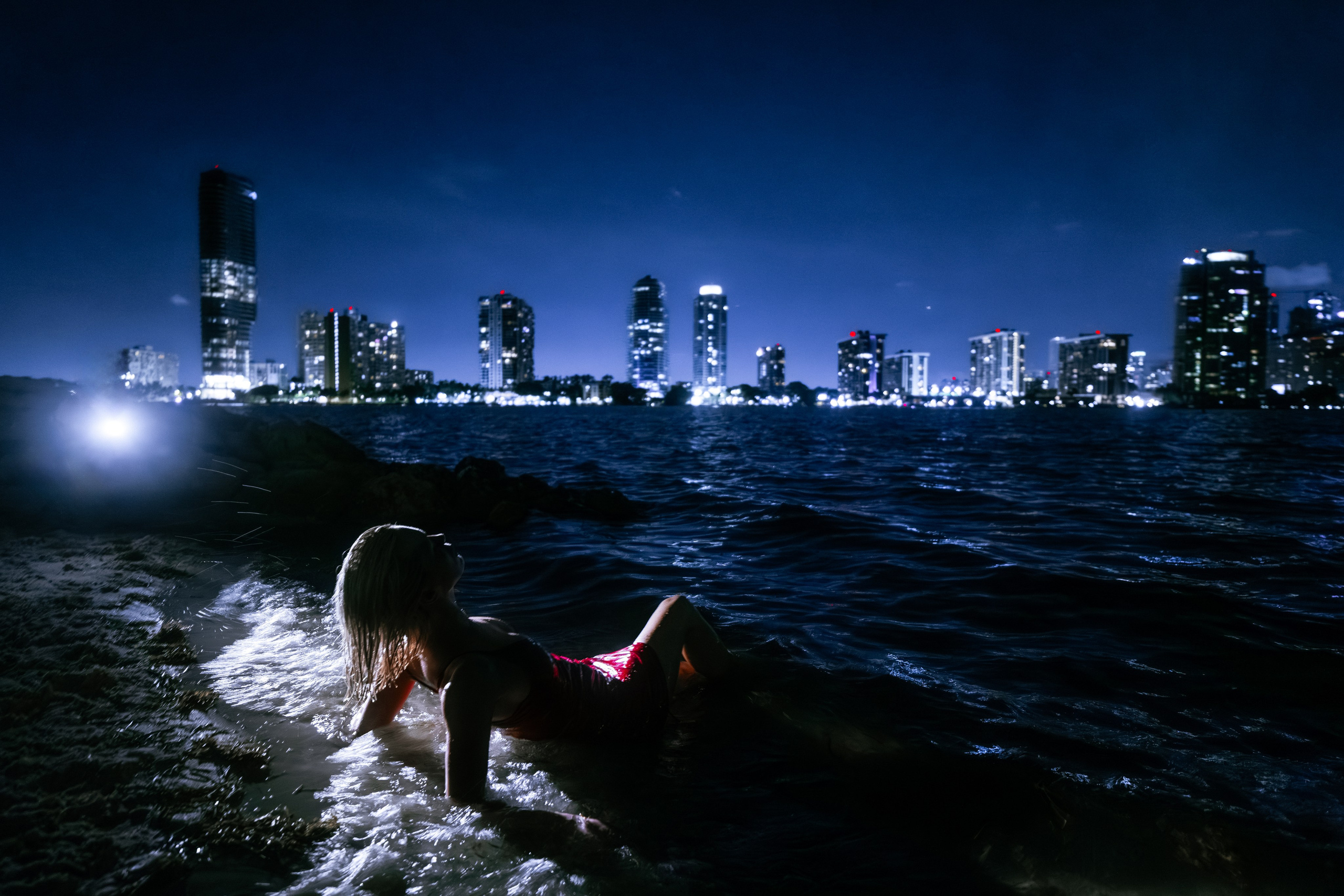 Reclining pose on the shoreline, the body aligns with the horizon, the red dress forming a striking accent against the night. Shot with precise directional lighting. Stylish and minimal composition.