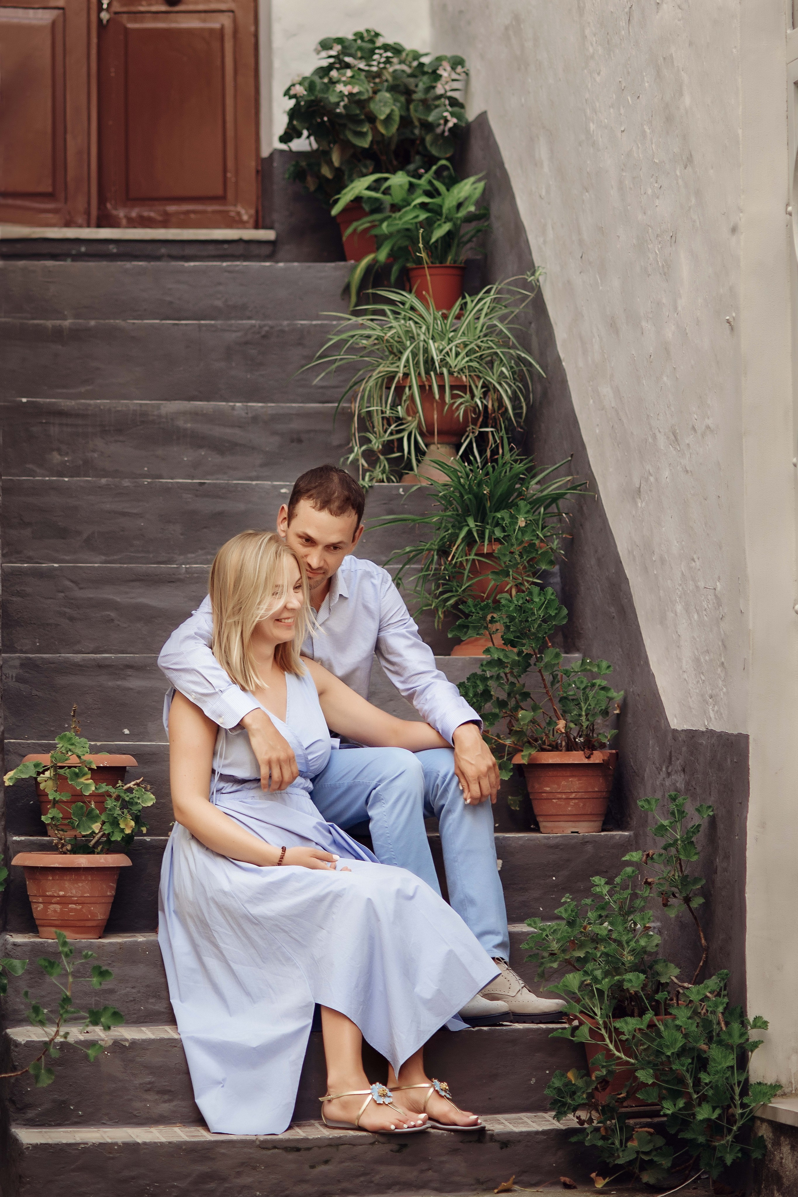 A couple sitting on stone steps surrounded by potted greenery. He gently hugs her shoulders.