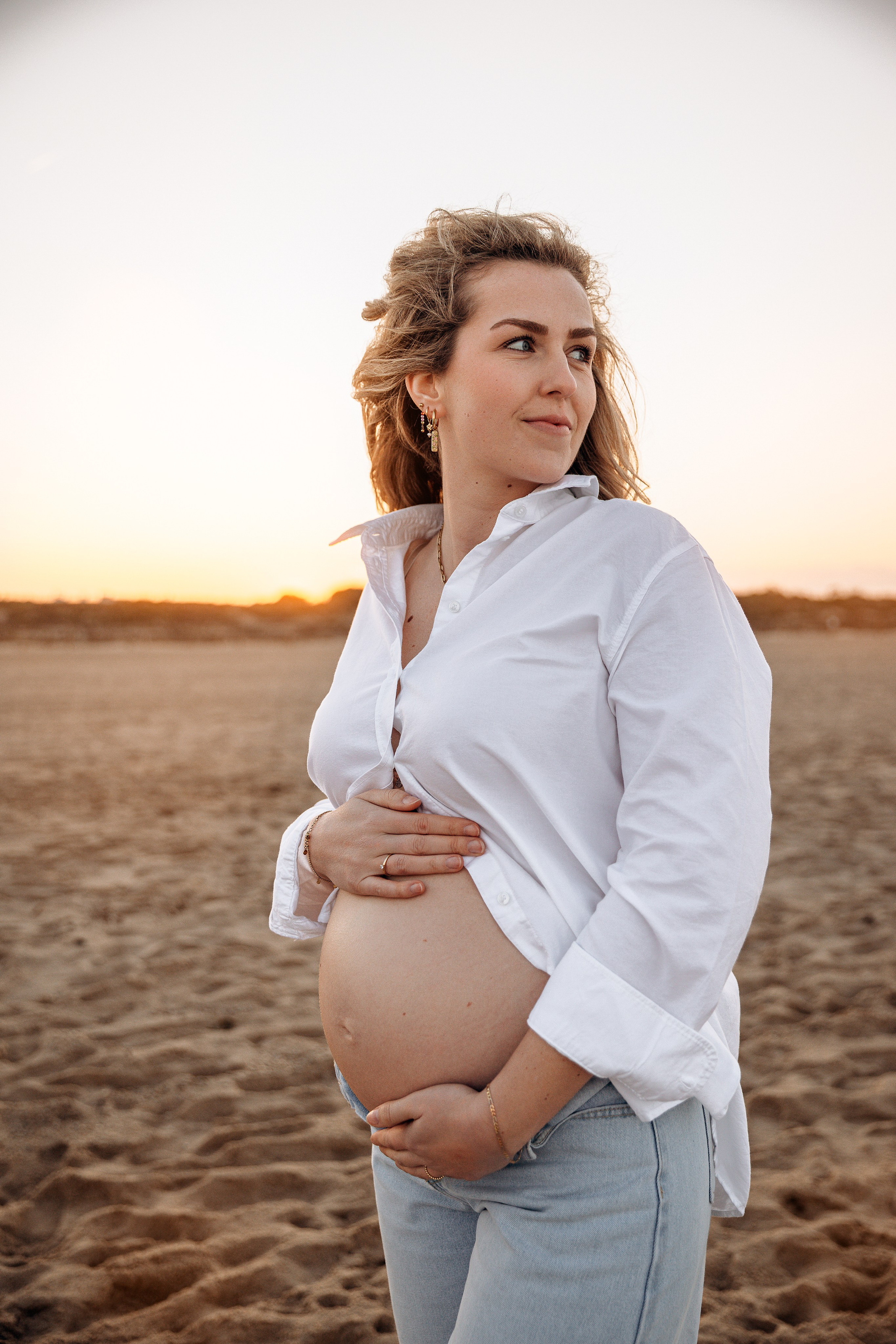 Sesión de fotos de embarazo al aire libre en Valencia, España, capturando a una mujer embarazada con una camisa blanca sosteniendo su barriga durante la hora dorada.