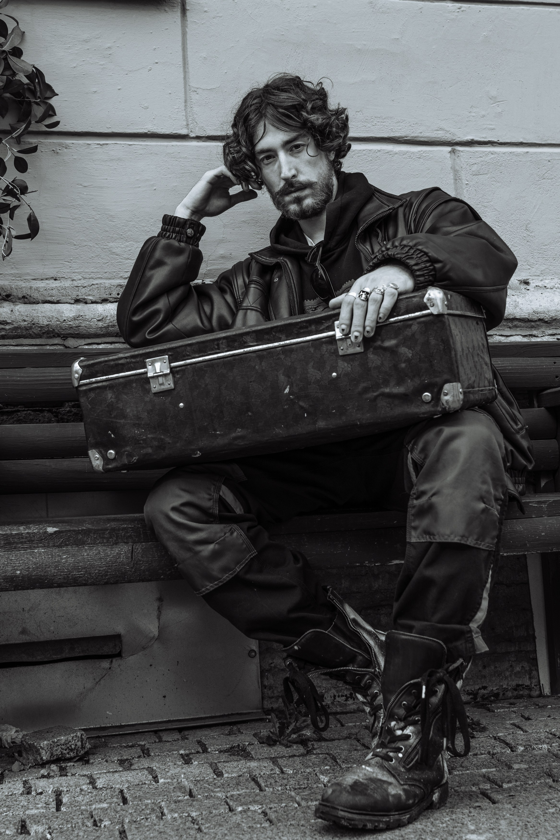 Мужчина с волосами до плеч сидит на скамейке и держит на коленях чемодан