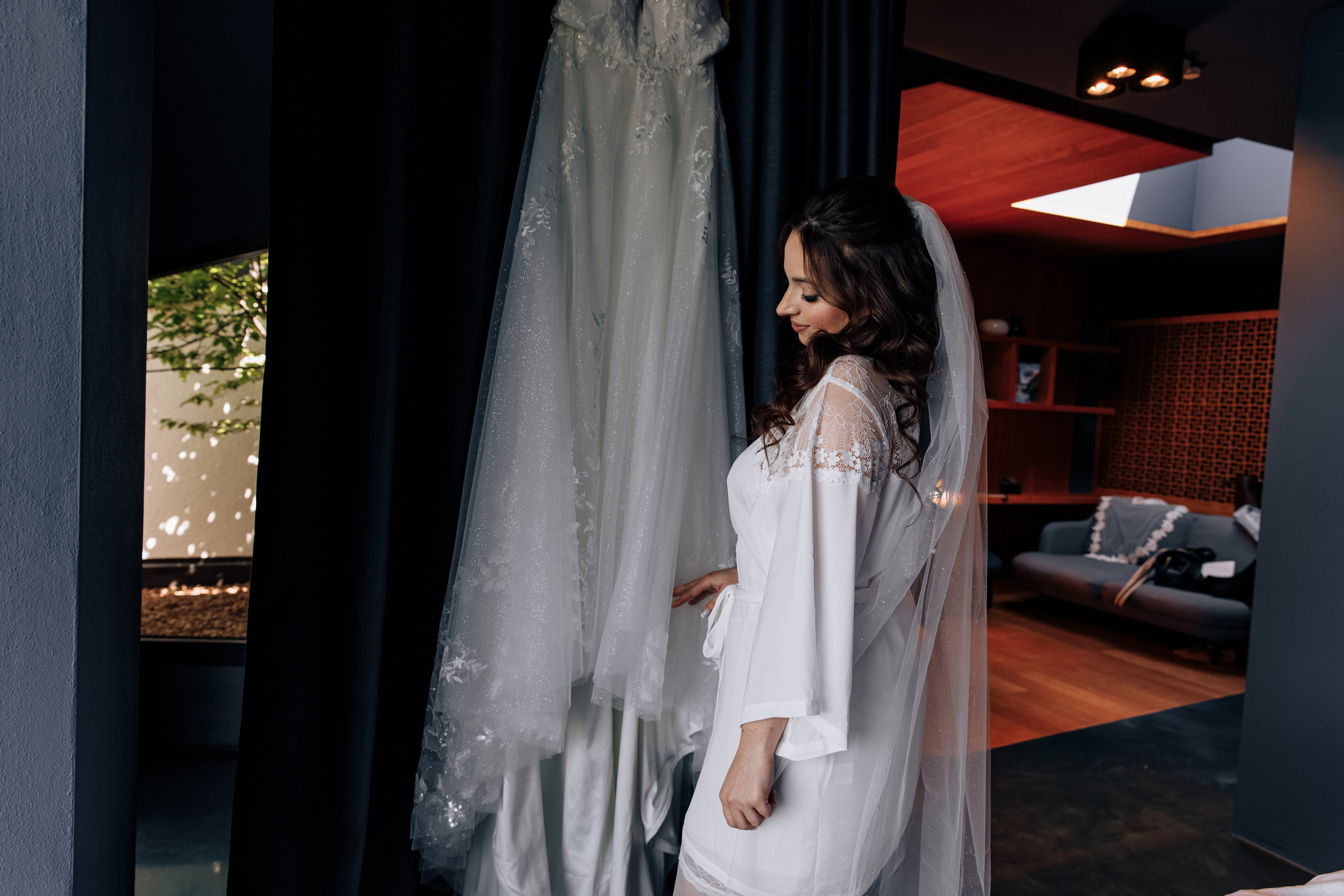 Die Braut streicht sanft über den Saum ihres Hochzeitskleides, das noch an der Wand hängt. 